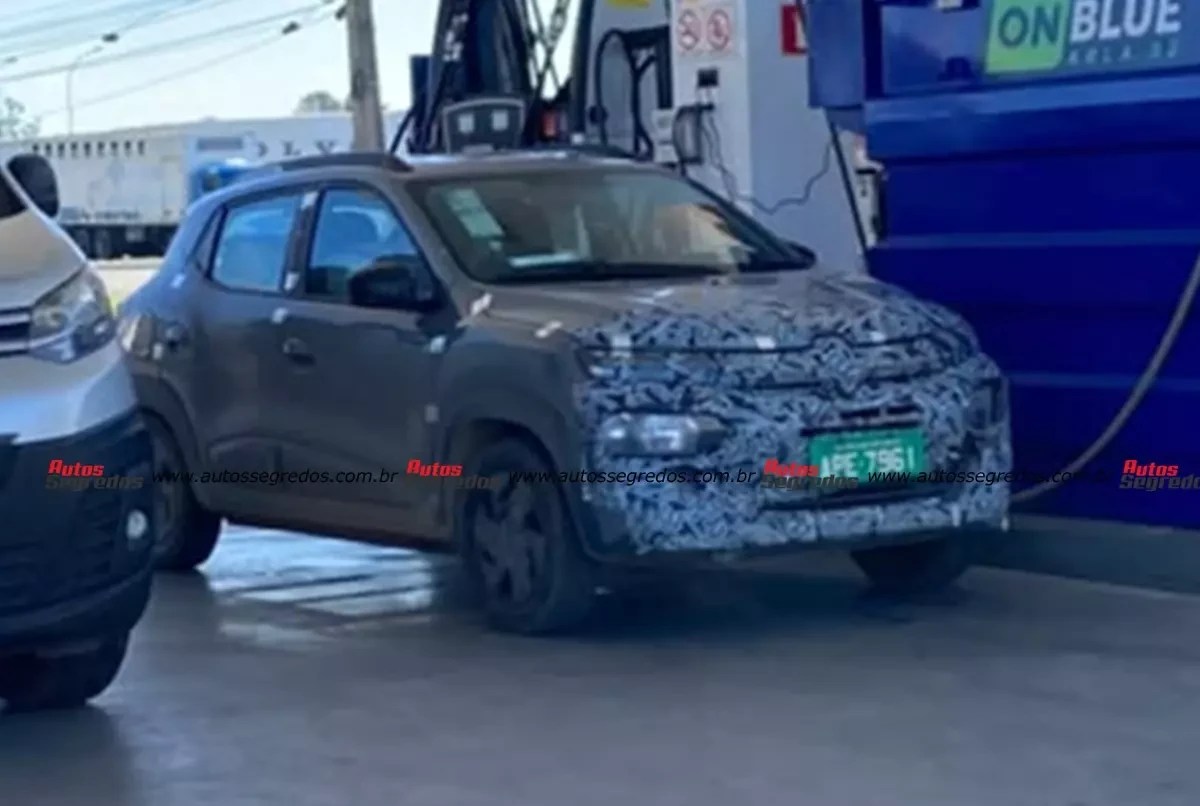 Carro cinza escuro camuflado com adesivos azuis e brancos, placa verde APE-7961, parado em posto de gasolina ao lado de bomba azul com ON BLUE escrito. Outro carro prateado aparece parcialmente à esquerda