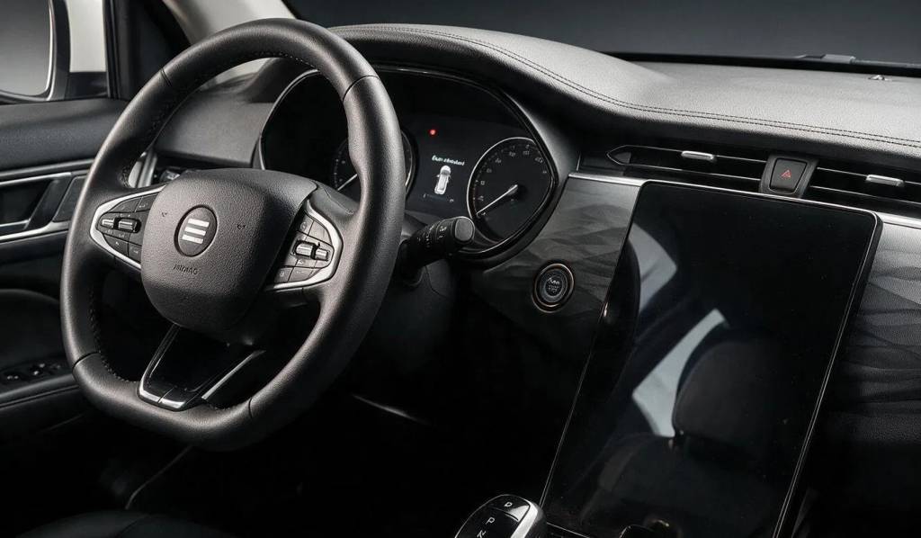 Interior de um carro moderno com volante multifuncional de couro preto, painel digital com veloc&iacute;metro e tela multim&iacute;dia vertical escura. Detalhes em cinza e preto texturizado no painel, bot&atilde;o de partida e alavanca de c&acirc;mbio autom&aacute;tica.