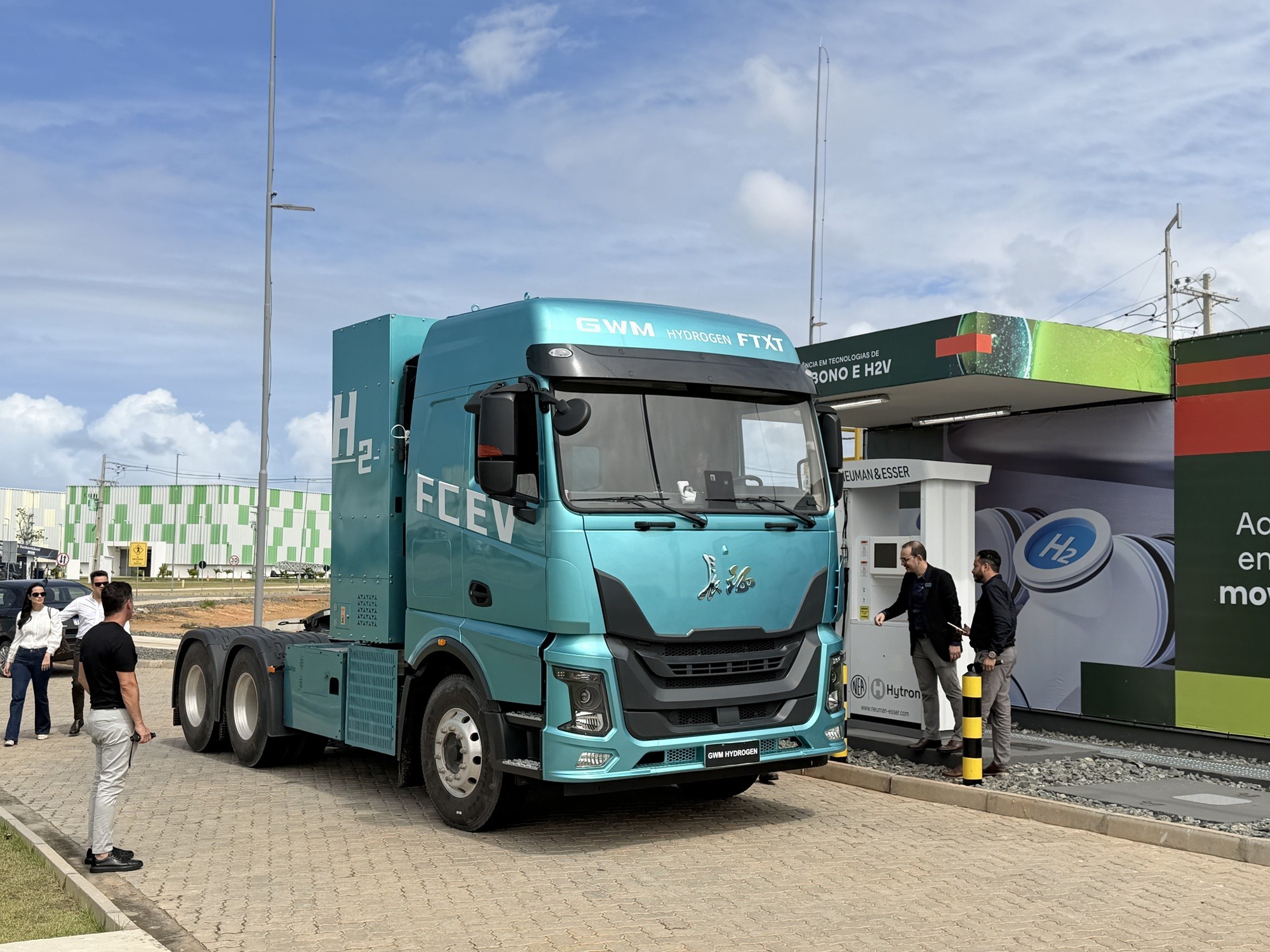 Caminh&atilde;o azul-turquesa com a inscri&ccedil;&atilde;o H2 FCEV na lateral, parado em um posto de abastecimento de hidrog&ecirc;nio. Dois homens observam o painel de uma bomba de combust&iacute;vel, enquanto outras pessoas conversam ao fundo.