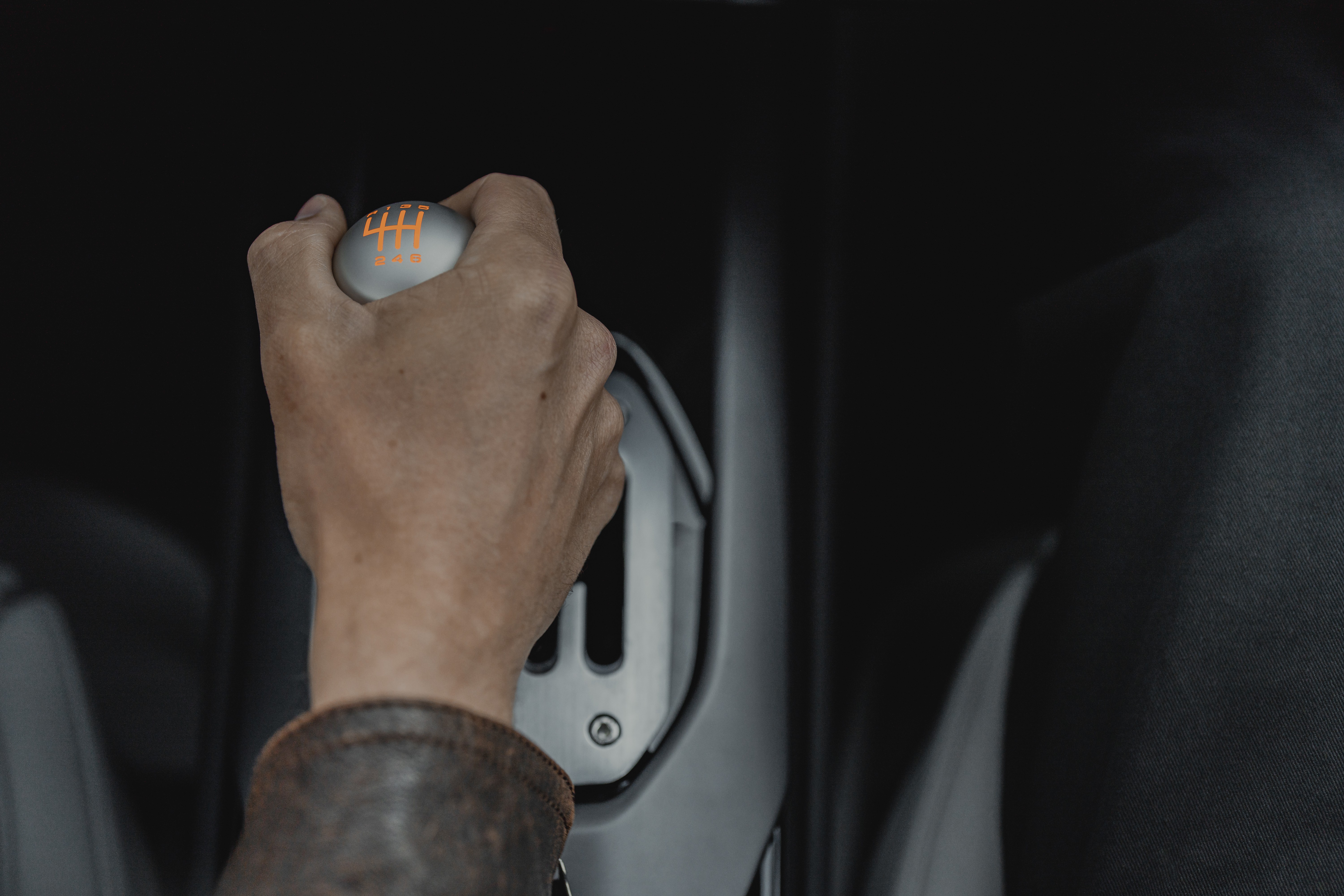 M&atilde;o de uma pessoa com jaqueta de couro marrom segurando o c&acirc;mbio manual de um carro, com a manopla cinza e n&uacute;meros de marcha laranja, em um interior escuro