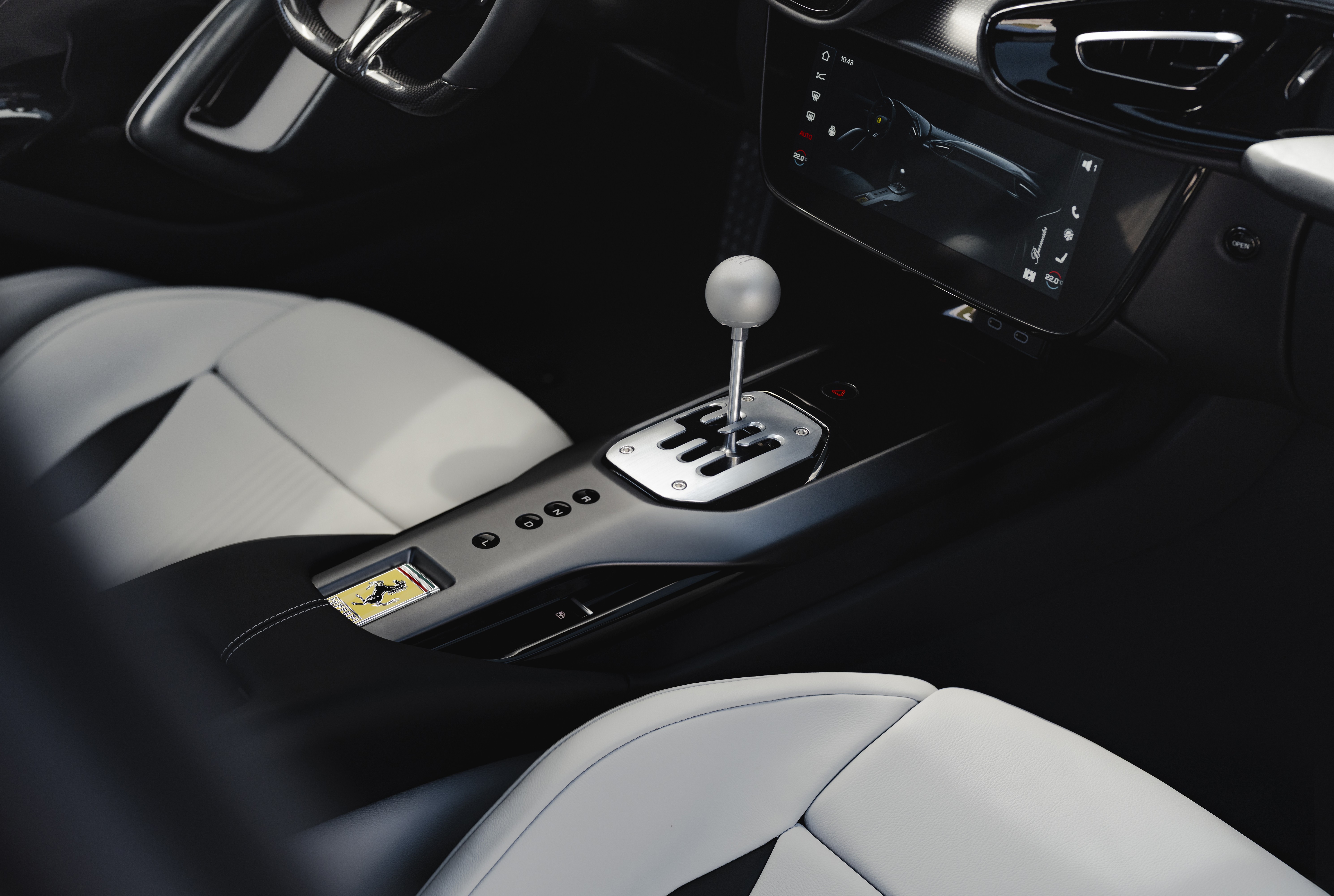Interior de um carro esportivo com bancos de couro branco e preto, console central cinza escuro e c&acirc;mbio manual cromado com manopla esf&eacute;rica. O painel possui uma tela multim&iacute;dia e o volante &eacute; vis&iacute;vel no canto superior esquerdo. No console, h&aacute; um emblema dourado da Ferrari