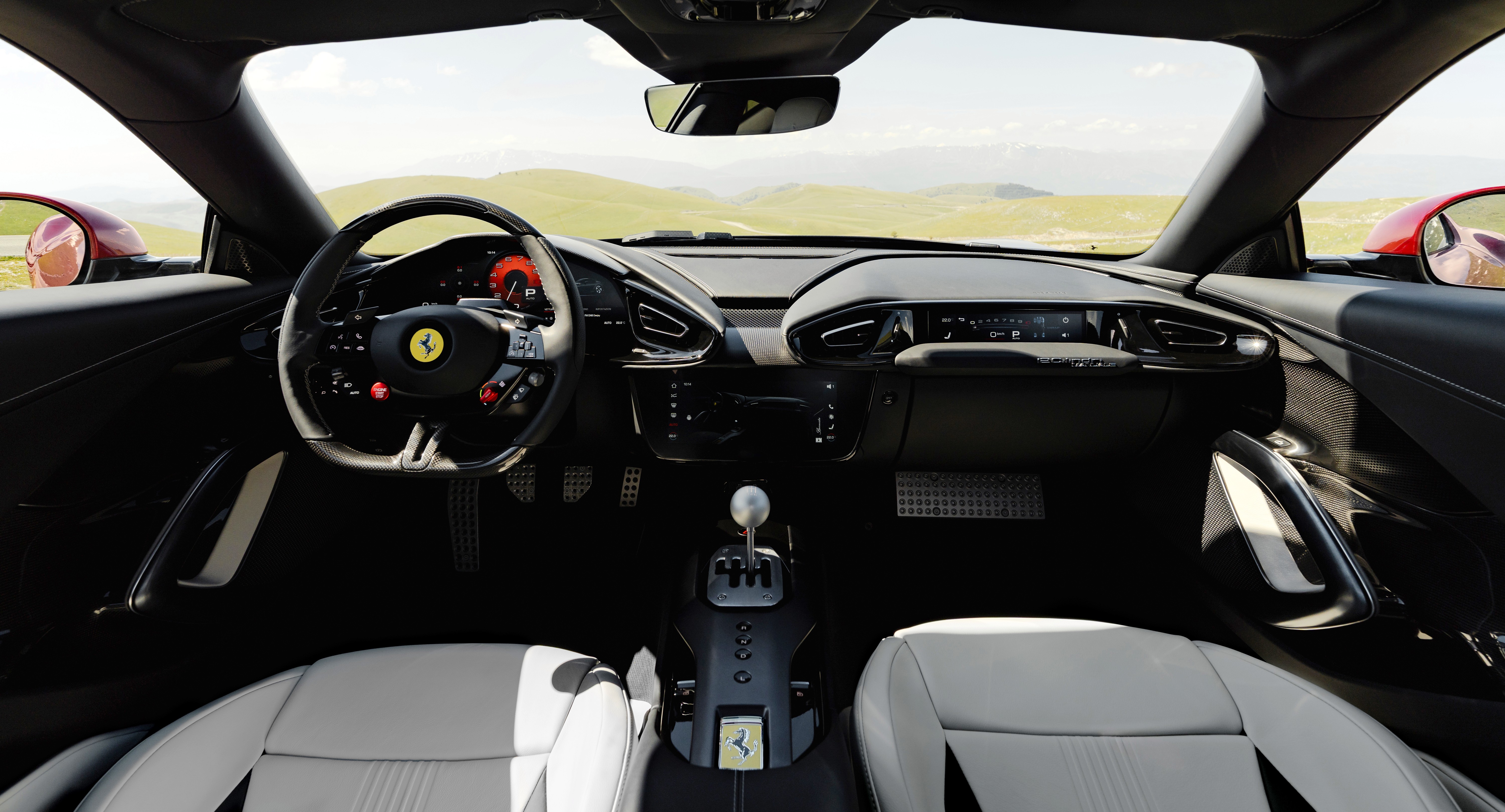 Interior de um carro esportivo Ferrari, com bancos de couro branco, volante preto com o emblema da Ferrari e detalhes vermelhos. O painel &eacute; escuro, com um c&acirc;mbio manual prateado no console central. Ao fundo, uma paisagem montanhosa verde sob c&eacute;u claro, vis&iacute;vel atrav&eacute;s do para-brisa e dos retrovisores laterais vermelhos