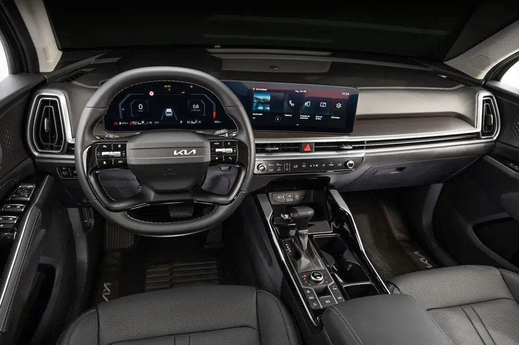 Interior moderno de um carro, com volante multifuncional preto e logo Kia. Painel digital exibe informa&ccedil;&otilde;es do ve&iacute;culo e uma tela multim&iacute;dia sens&iacute;vel ao toque mostra &iacute;cones de aplicativos. O console central possui alavanca de c&acirc;mbio e bot&otilde;es de controle, bancos de couro escuro e detalhes prateados no painel. A ilumina&ccedil;&atilde;o &eacute; suave, destacando o design sofisticado
