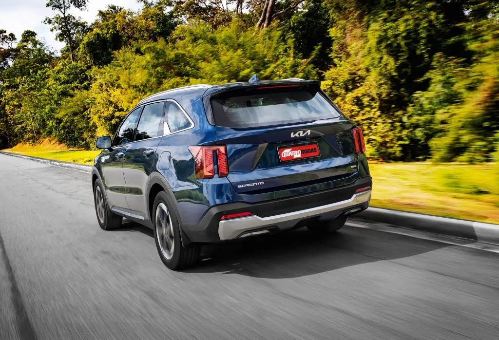 SUV Kia Sorento azul escuro visto por tr&aacute;s, em movimento numa estrada asfaltada, com &aacute;rvores e vegeta&ccedil;&atilde;o verde ao fundo