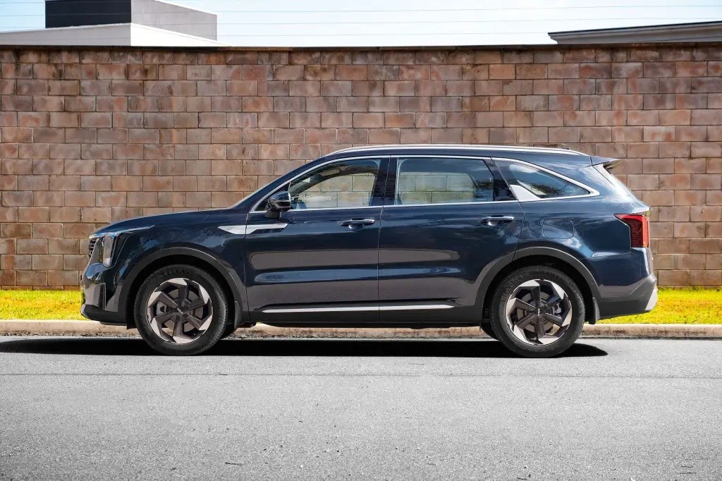 SUV Kia Sorento azul-escuro estacionado de perfil em uma rua asfaltada, com uma parede de tijolos e grama verde ao fundo