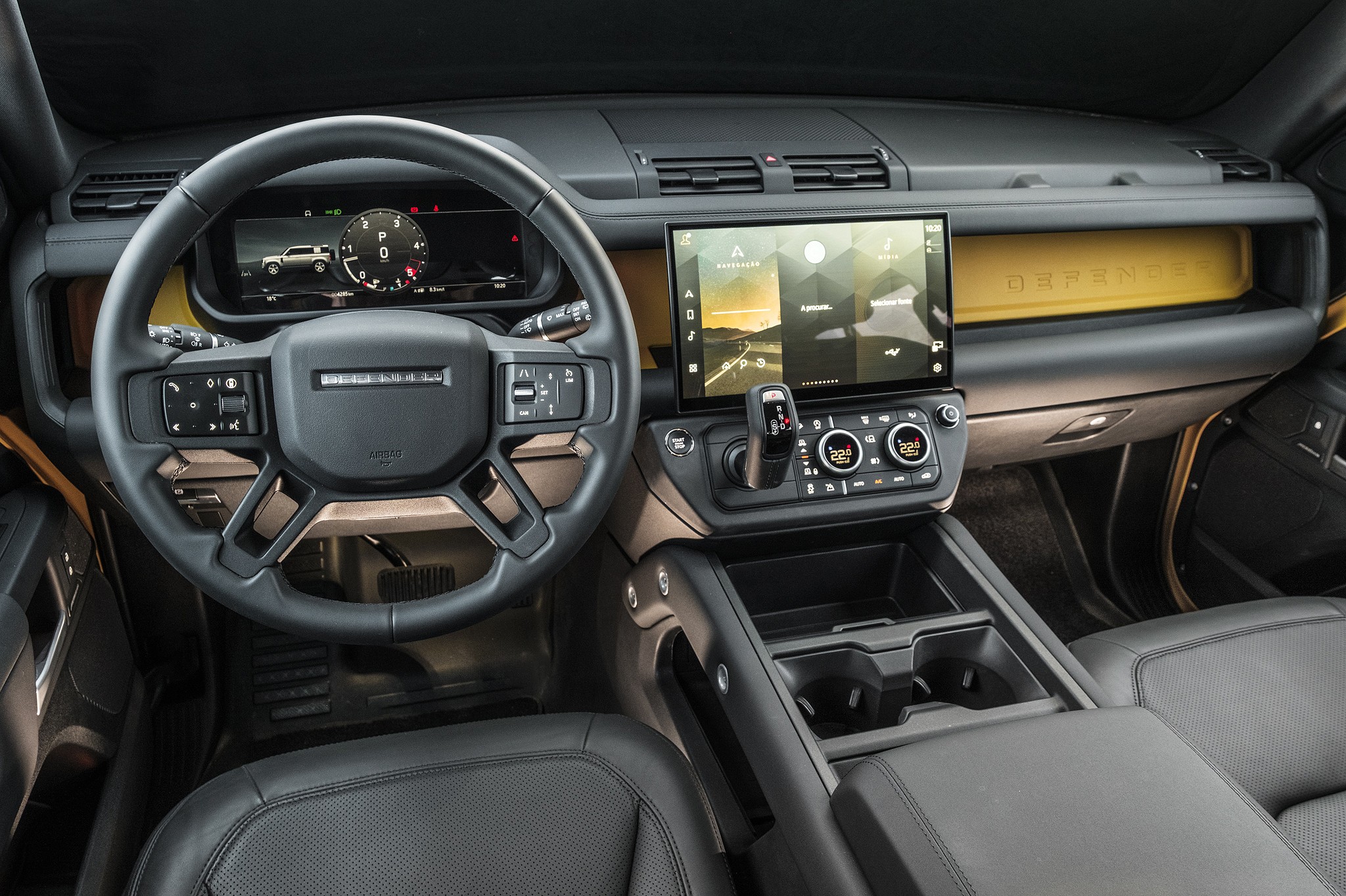 Interior de um carro moderno com volante multifuncional preto, painel digital exibindo um ve&iacute;culo e um veloc&iacute;metro, e uma tela multim&iacute;dia central com paisagem. O painel tem detalhes em amarelo e o console central possui porta-copos e apoio de bra&ccedil;o. Os bancos s&atilde;o de couro preto.