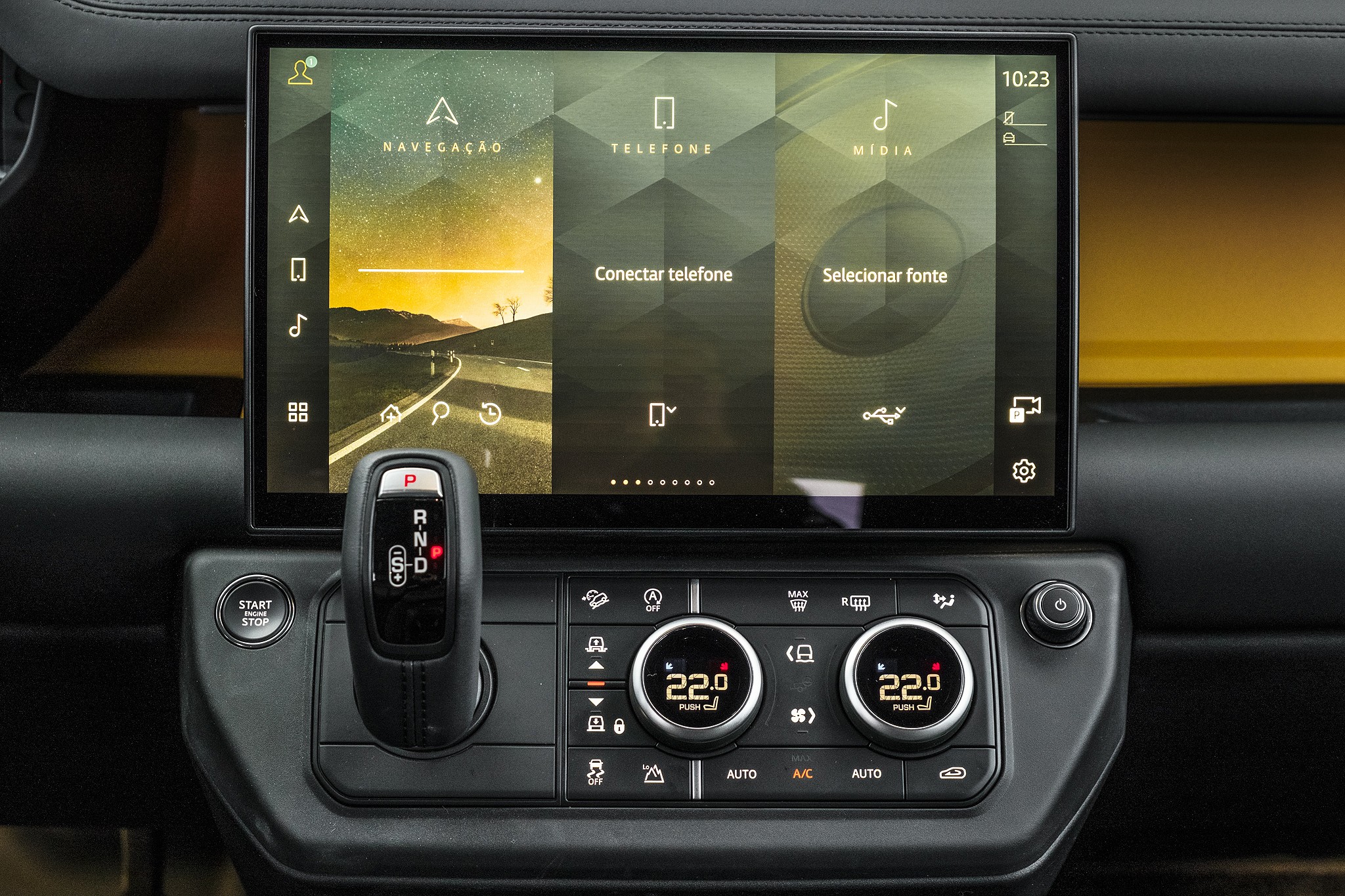 Painel interno de um carro com tela multim&iacute;dia exibindo op&ccedil;&otilde;es de navega&ccedil;&atilde;o, telefone e m&iacute;dia, com uma estrada ao p&ocirc;r do sol na tela de navega&ccedil;&atilde;o. Abaixo, alavanca de c&acirc;mbio, bot&atilde;o Start Stop e controles de ar condicionado com temperatura de 22 graus