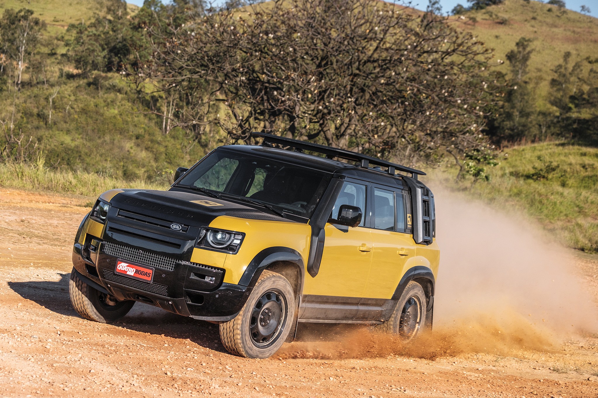 SUV Land Rover Defender amarelo e preto em estrada de terra, levantando poeira. O ve&iacute;culo tem rack de teto e snorkel, com colinas verdes e &aacute;rvores ao fundo