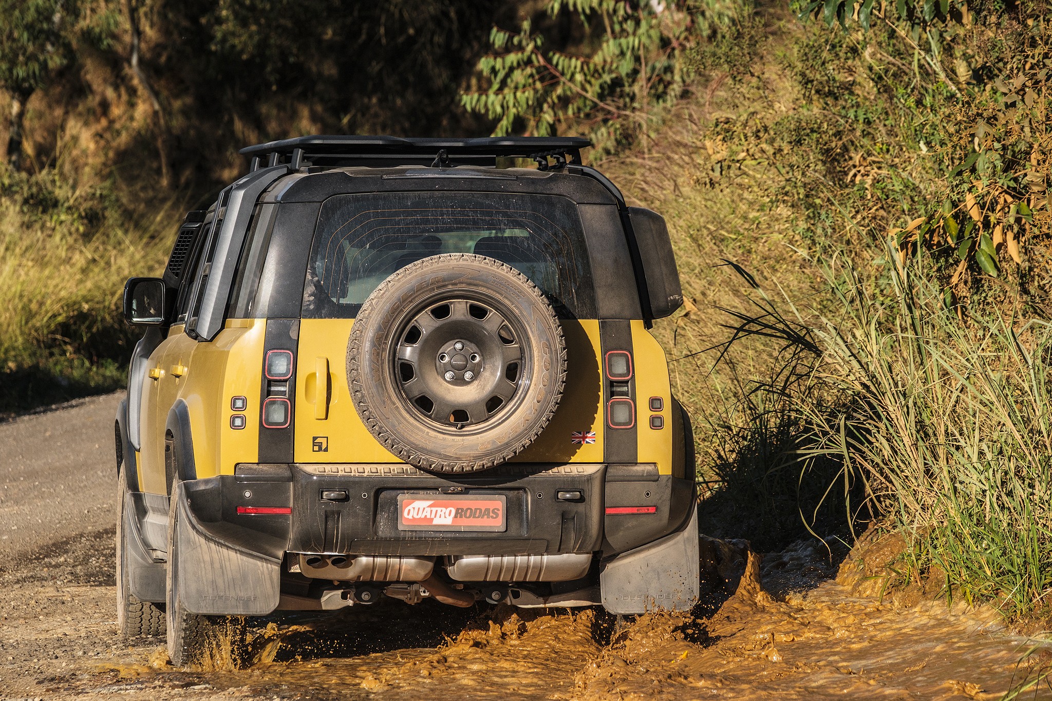 Traseira de um Land Rover Defender amarelo e preto em estrada de terra, com lama espirrando dos pneus. O carro tem estepe na porta traseira, rack de teto e para-barros. Vegeta&ccedil;&atilde;o seca e verde nas laterais da estrada, sob luz solar