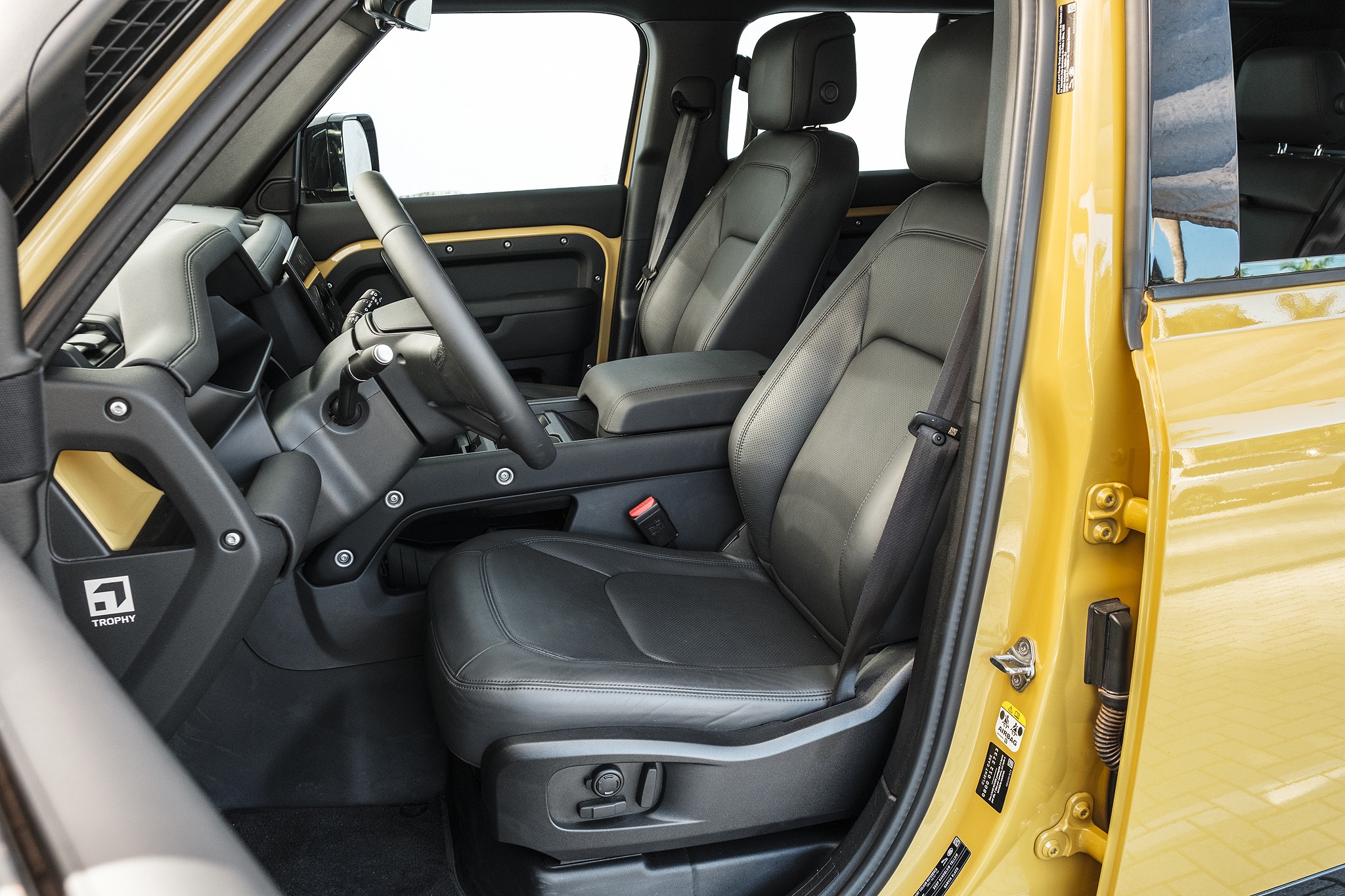 Interior de um carro amarelo com bancos de couro preto, volante e painel escuros. O banco do motorista est&aacute; em primeiro plano, com cinto de seguran&ccedil;a vis&iacute;vel. Detalhes dourados no painel e na porta contrastam com o preto, e um emblema Trophy aparece na lateral do console central
