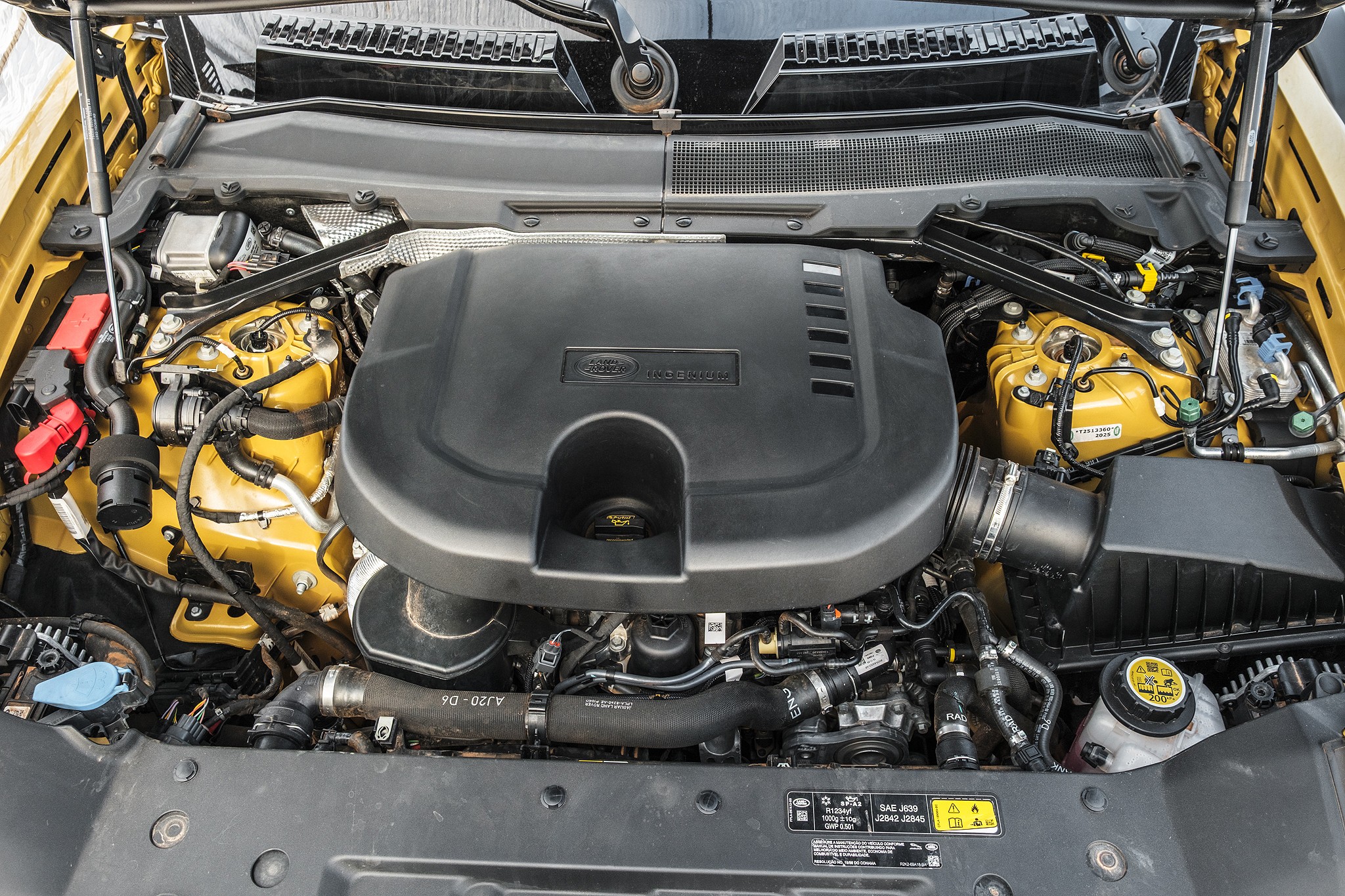 Vista superior do motor de um carro, com a tampa preta do motor em destaque no centro, cercada por componentes amarelos e mangueiras pretas. O cap&ocirc; est&aacute; aberto, revelando a complexidade do compartimento do motor, com detalhes como o reservat&oacute;rio de fluido de freio e o filtro de ar vis&iacute;veis. O fundo &eacute; um tom de amarelo, indicando a cor do ve&iacute;culo