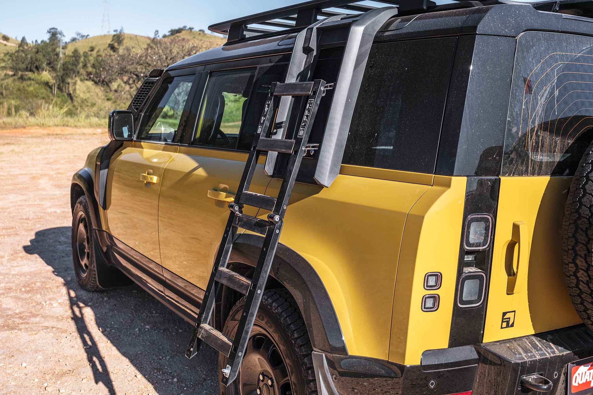Lateral e traseira de um SUV amarelo com teto preto, rack de teto e escada preta dobr&aacute;vel na lateral. O ve&iacute;culo est&aacute; estacionado em uma estrada de terra, com colinas e vegeta&ccedil;&atilde;o ao fundo sob um c&eacute;u azul