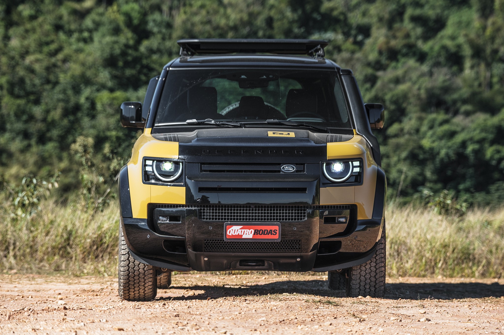 Land Rover Defender amarelo e preto, com bagageiro de teto, parado em estrada de terra. Far&oacute;is acesos, grade frontal preta com o logo da Land Rover e placa Quatro Rodas vermelha. Ao fundo, vegeta&ccedil;&atilde;o verde e seca