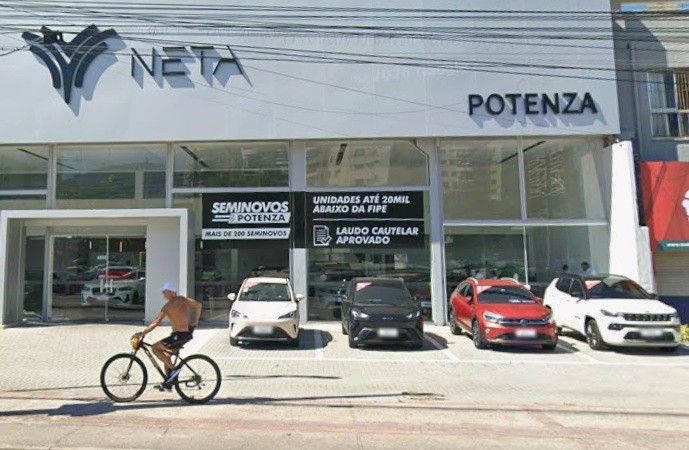 Homem sem camisa e de bon&eacute; branco pedala uma bicicleta preta em frente &agrave; concession&aacute;ria Neta Potenza, com carros expostos e an&uacute;ncios de seminovos e laudo cautelar aprovado