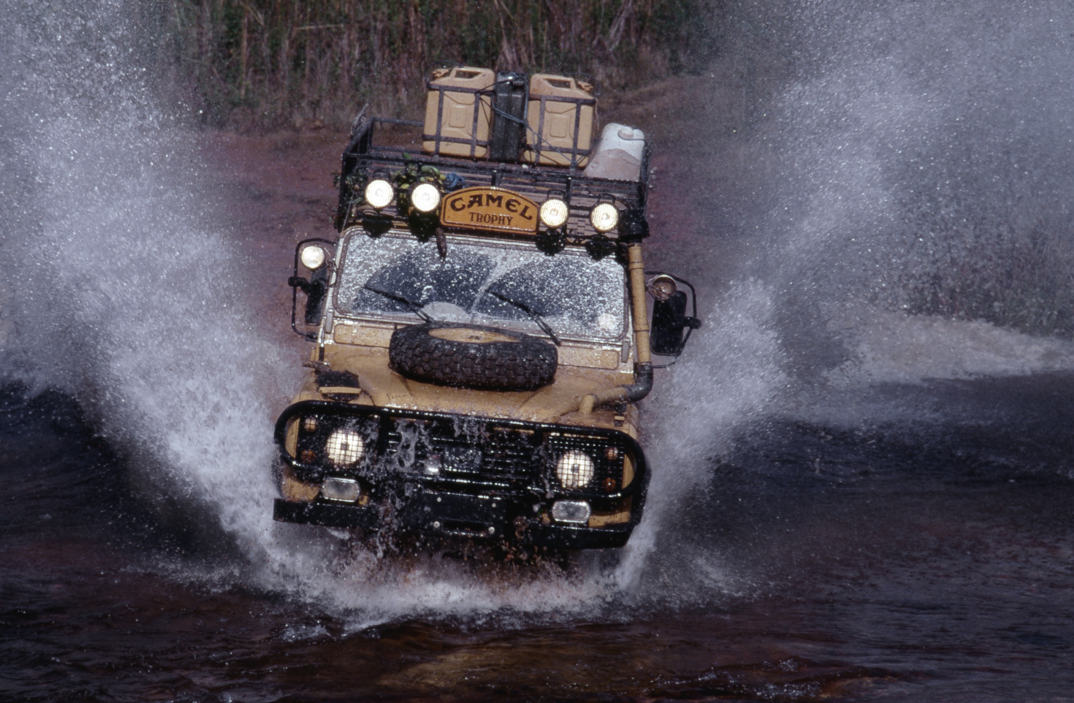 Jipe Land Rover Defender bege com Camel Trophy no teto, atravessando um rio, levantando grandes jatos de &aacute;gua para os lados e sobre o para-brisa
