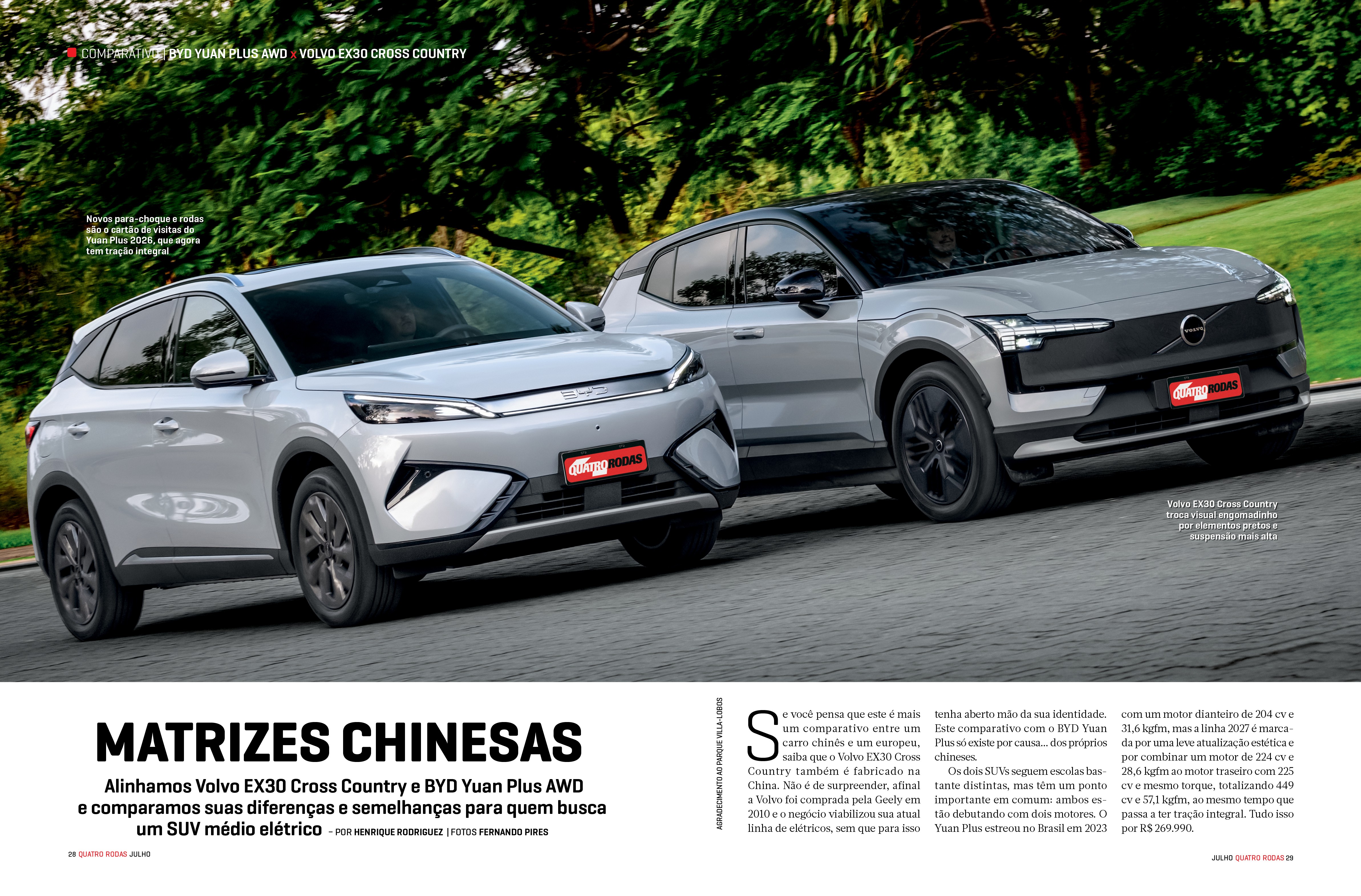 Dois SUVs el&eacute;tricos, um branco e um cinza escuro, lado a lado em uma estrada asfaltada, com &aacute;rvores verdes ao fundo