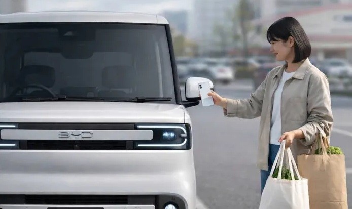 Mulher sorridente, com cabelo curto e jaqueta bege, destrava um carro branco da BYD usando o celular no retrovisor, segurando sacolas de compras