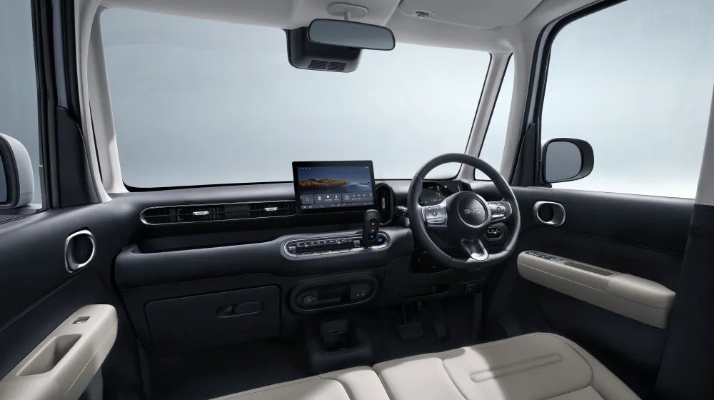 Interior de um carro moderno, com painel escuro e detalhes claros. Volante multifuncional &agrave; direita, tela multim&iacute;dia central exibindo uma paisagem. Bancos de couro bege e espelho retrovisor vis&iacute;vel.