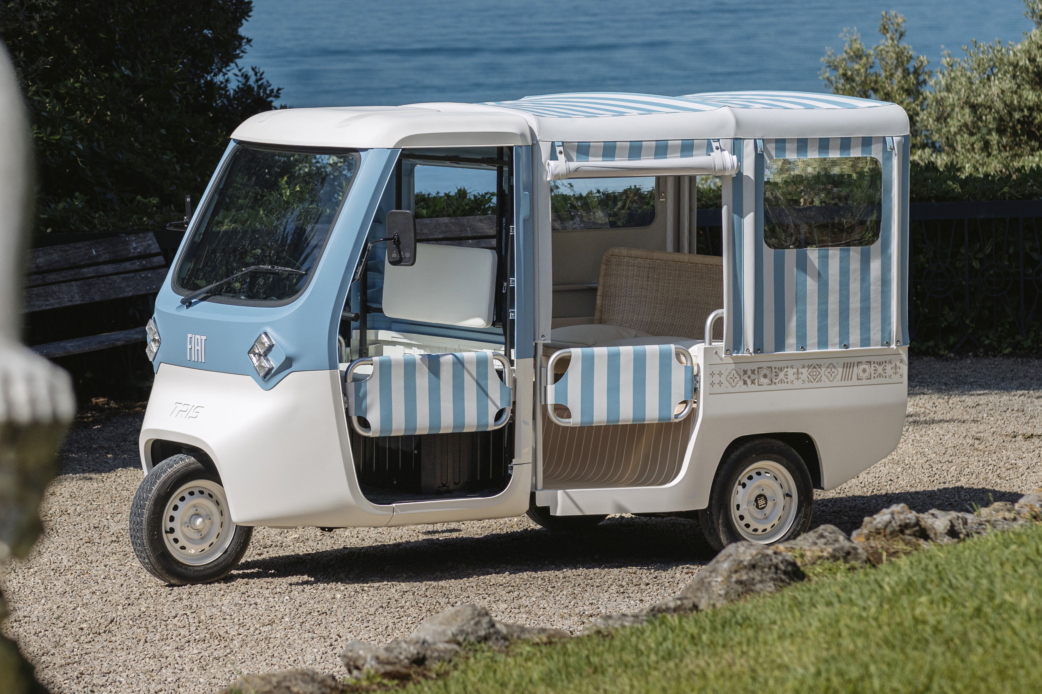 Um ve&iacute;culo el&eacute;trico de tr&ecirc;s rodas, branco e azul-claro, com teto e laterais listrados em azul e branco, estacionado em um caminho de cascalho. O mar azul e a vegeta&ccedil;&atilde;o verde aparecem ao fundo