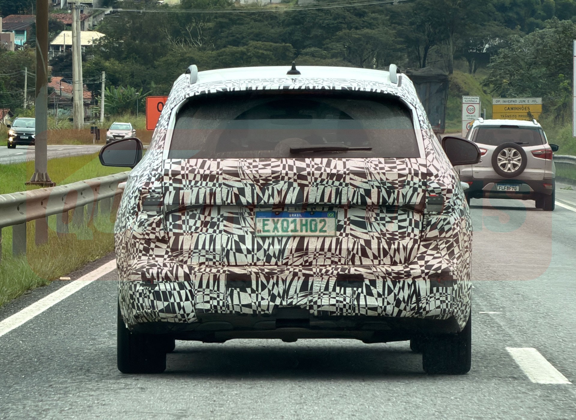 Traseira de um carro camuflado com adesivos pretos e brancos, placa EXQ1H02, em uma estrada. &Agrave; direita, um SUV prata com estepe na tampa traseira.
