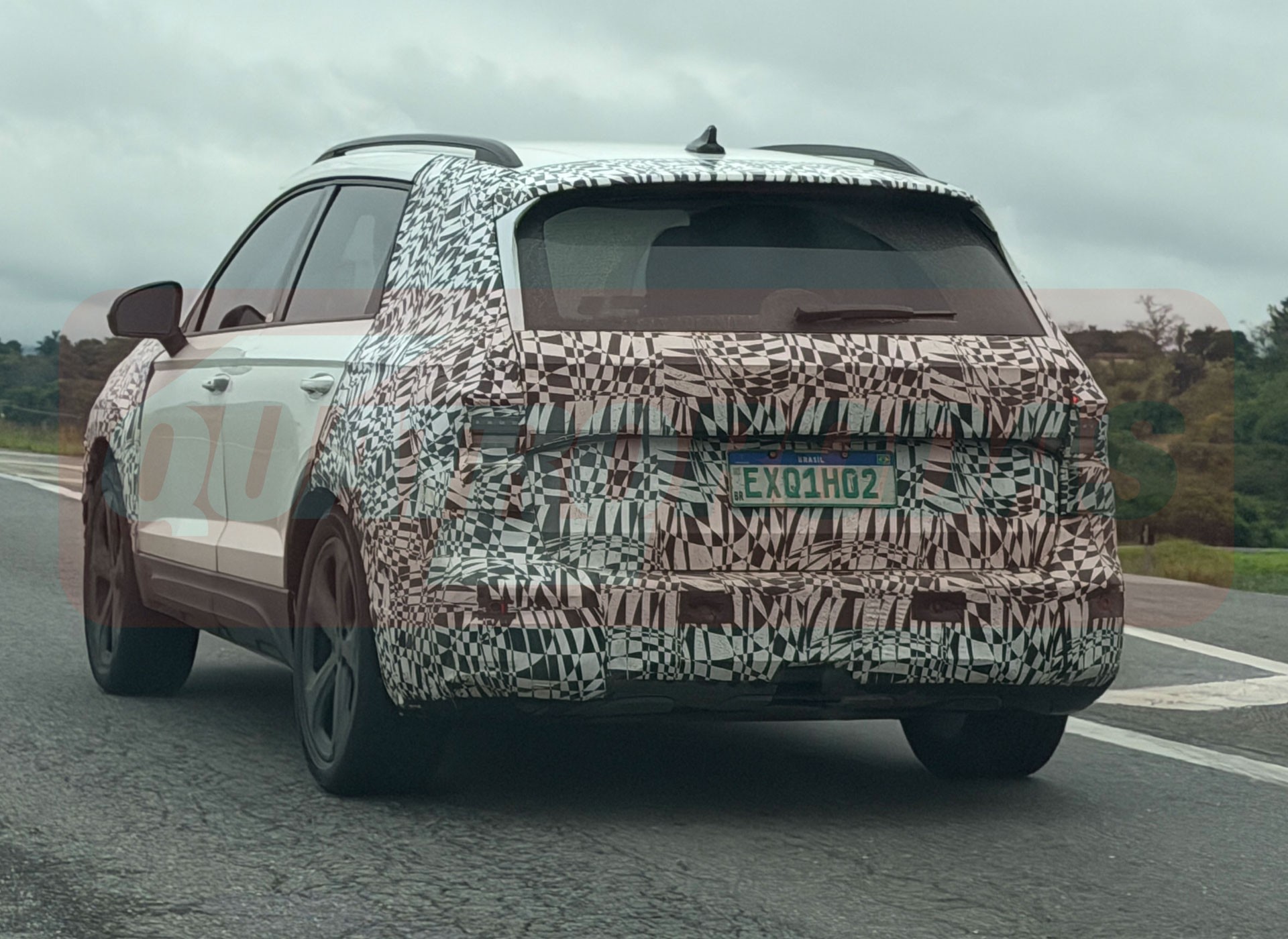 SUV branco camuflado com adesivos pretos e brancos, visto de traseira e lateral, trafegando em estrada sob c&eacute;u nublado. A placa de teste &eacute; verde com letras brancas EXQ1H02