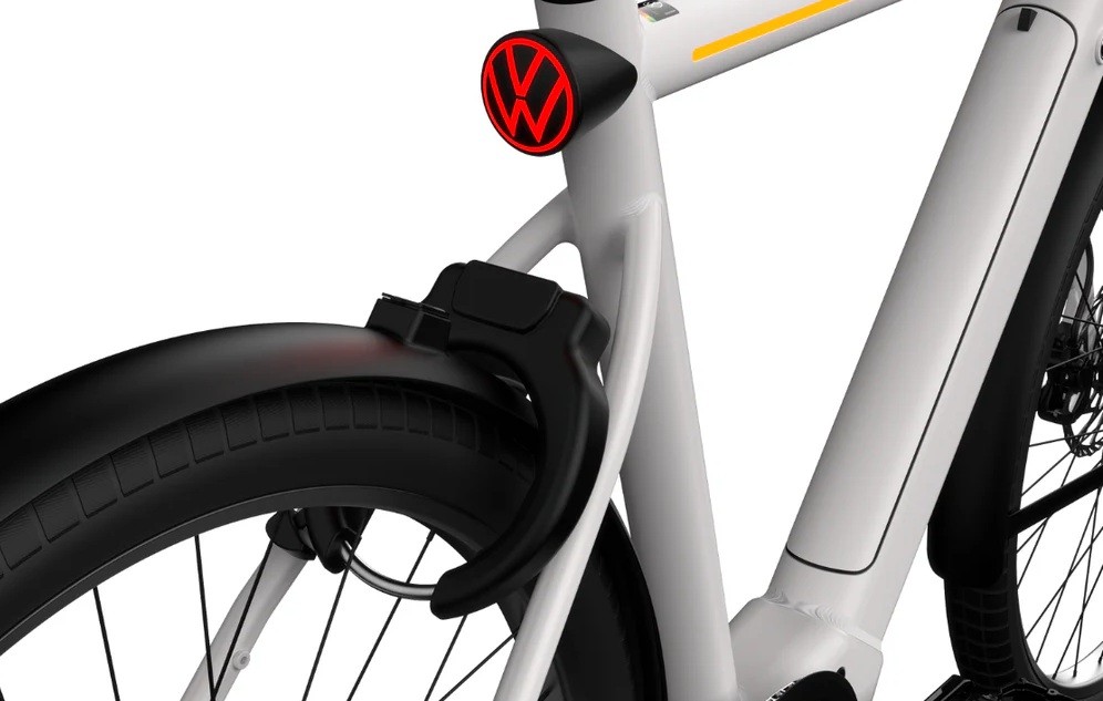 Detalhe da parte traseira de uma bicicleta el&eacute;trica branca, com para-lama preto e pneu com banda de rodagem. Acima do para-lama, uma lanterna traseira vermelha com o logotipo da Volkswagen em preto. Um cadeado em U preto est&aacute; preso ao quadro, pr&oacute;ximo &agrave; roda