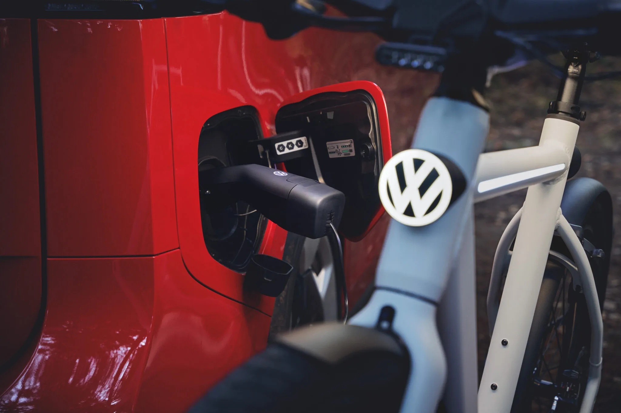 Carro vermelho sendo carregado por um cabo preto conectado &agrave; porta de carregamento, com uma bicicleta branca com o logo da Volkswagen em primeiro plano