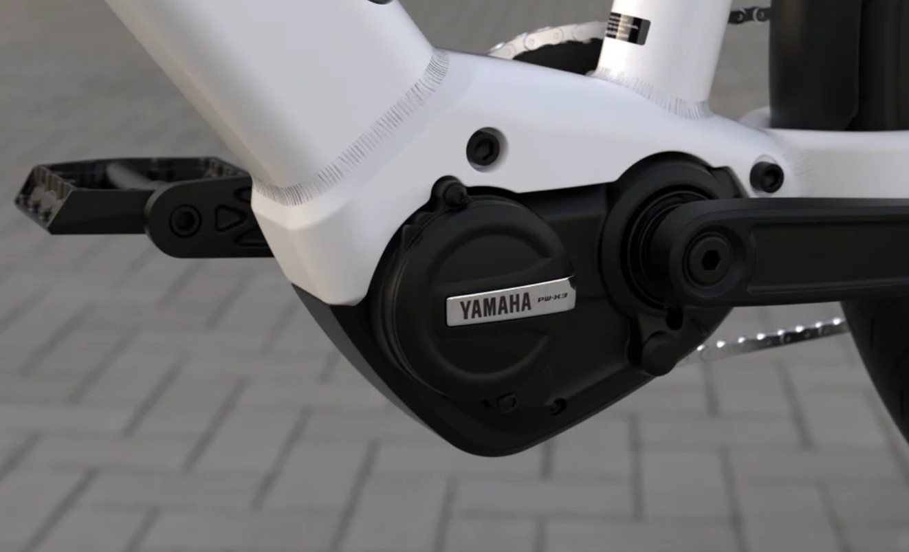 Motor el&eacute;trico Yamaha preto com logo prateado, integrado ao quadro branco de uma bicicleta, com pedal preto &agrave; esquerda, sobre piso de blocos cinzas