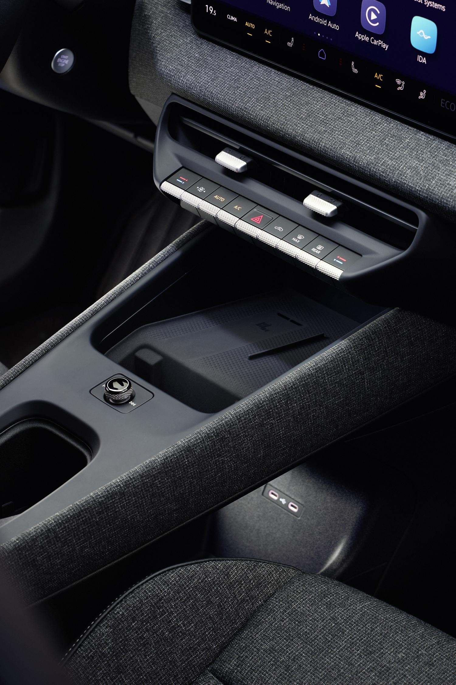 Interior de um carro moderno com painel cinza-escuro texturizado, tela multim&iacute;dia exibindo Android Auto e Apple CarPlay, controles de ar-condicionado e um carregador de celular por indu&ccedil;&atilde;o no console central
