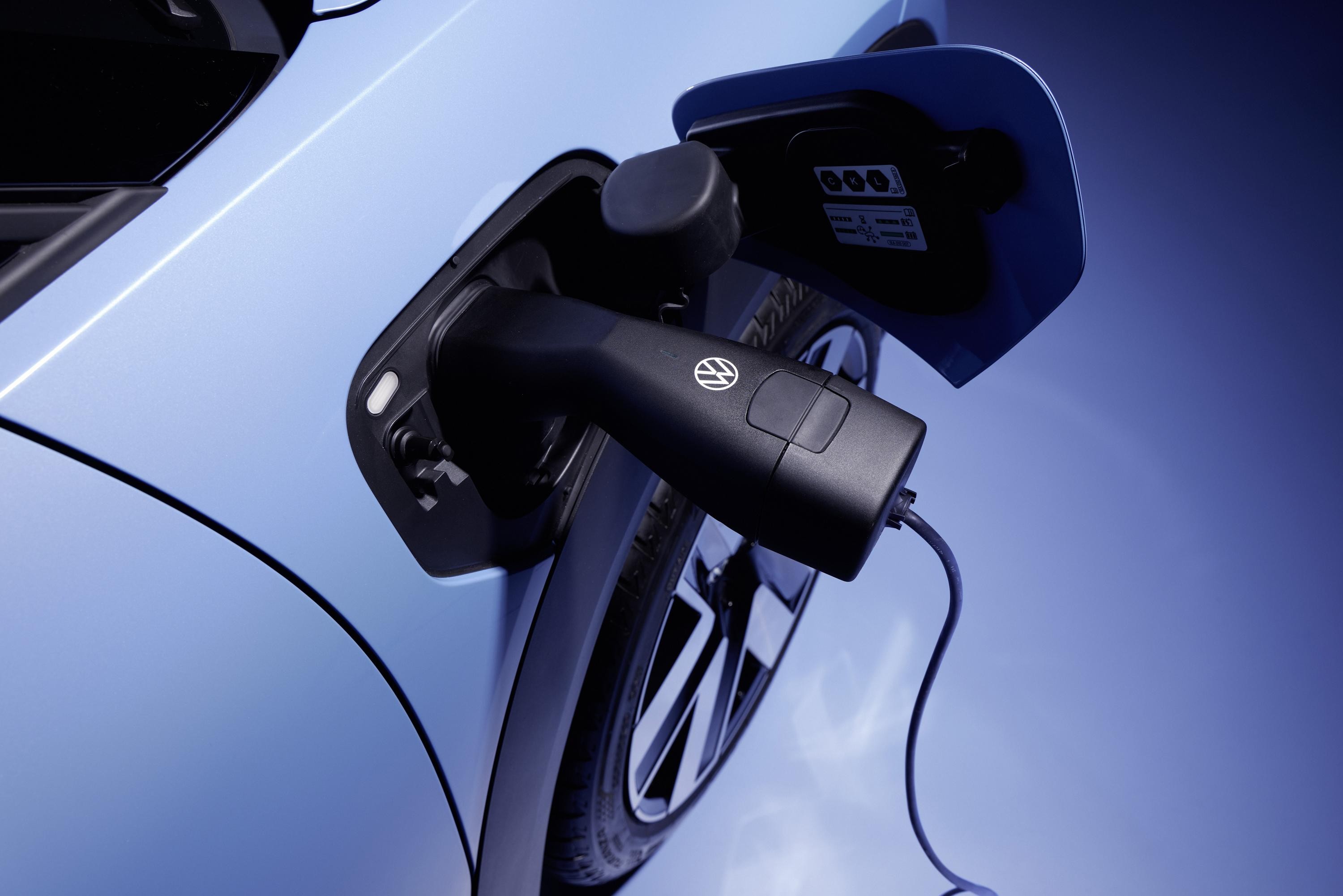 Carregador preto com logo Volkswagen conectado &agrave; entrada de um carro el&eacute;trico azul, com a tampa aberta e o cabo esticado