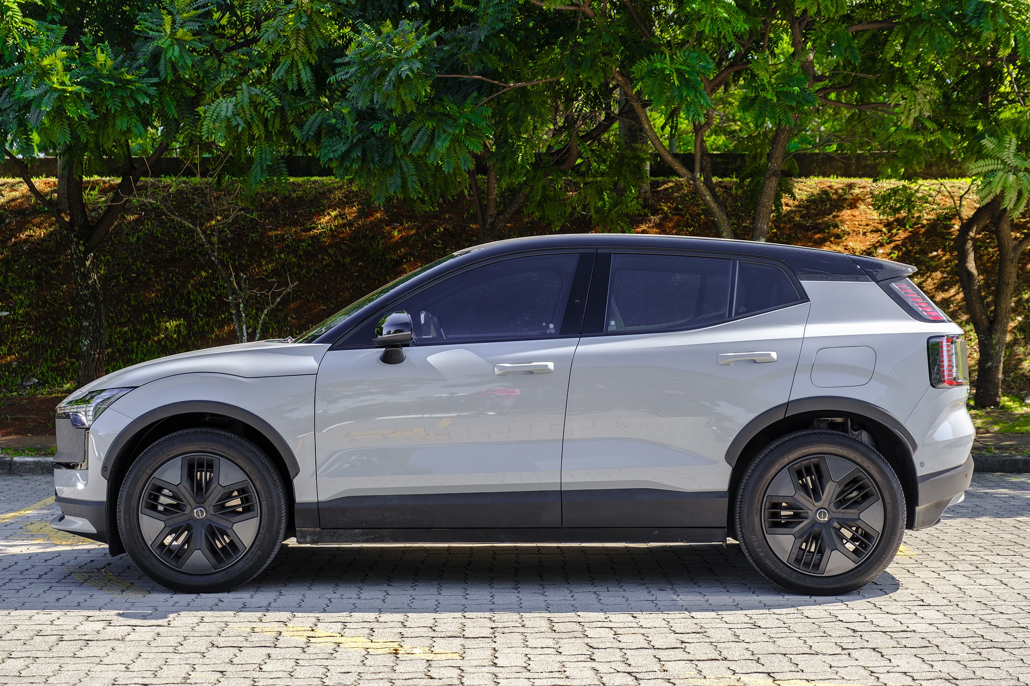Carro SUV el&eacute;trico cinza claro com teto preto, estacionado de lado em piso de paralelep&iacute;pedos, com &aacute;rvores verdes ao fundo