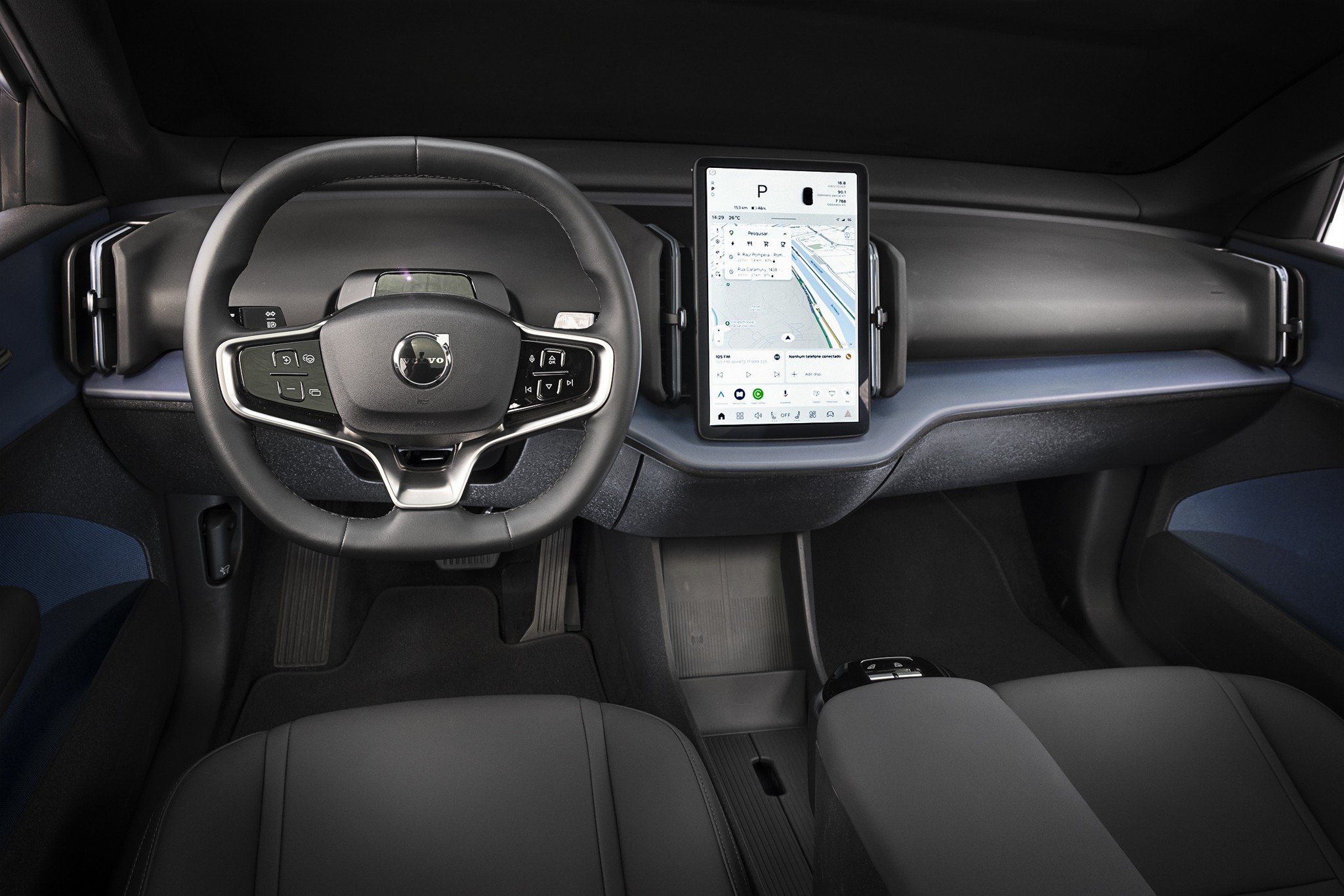 Interior de um carro moderno com volante preto e cinza, painel azul-acinzentado e tela vertical exibindo um mapa de navega&ccedil;&atilde;o. Os bancos dianteiros s&atilde;o pretos, e o console central &eacute; discreto, com um apoio de bra&ccedil;o. O ambiente &eacute; minimalista e tecnol&oacute;gico