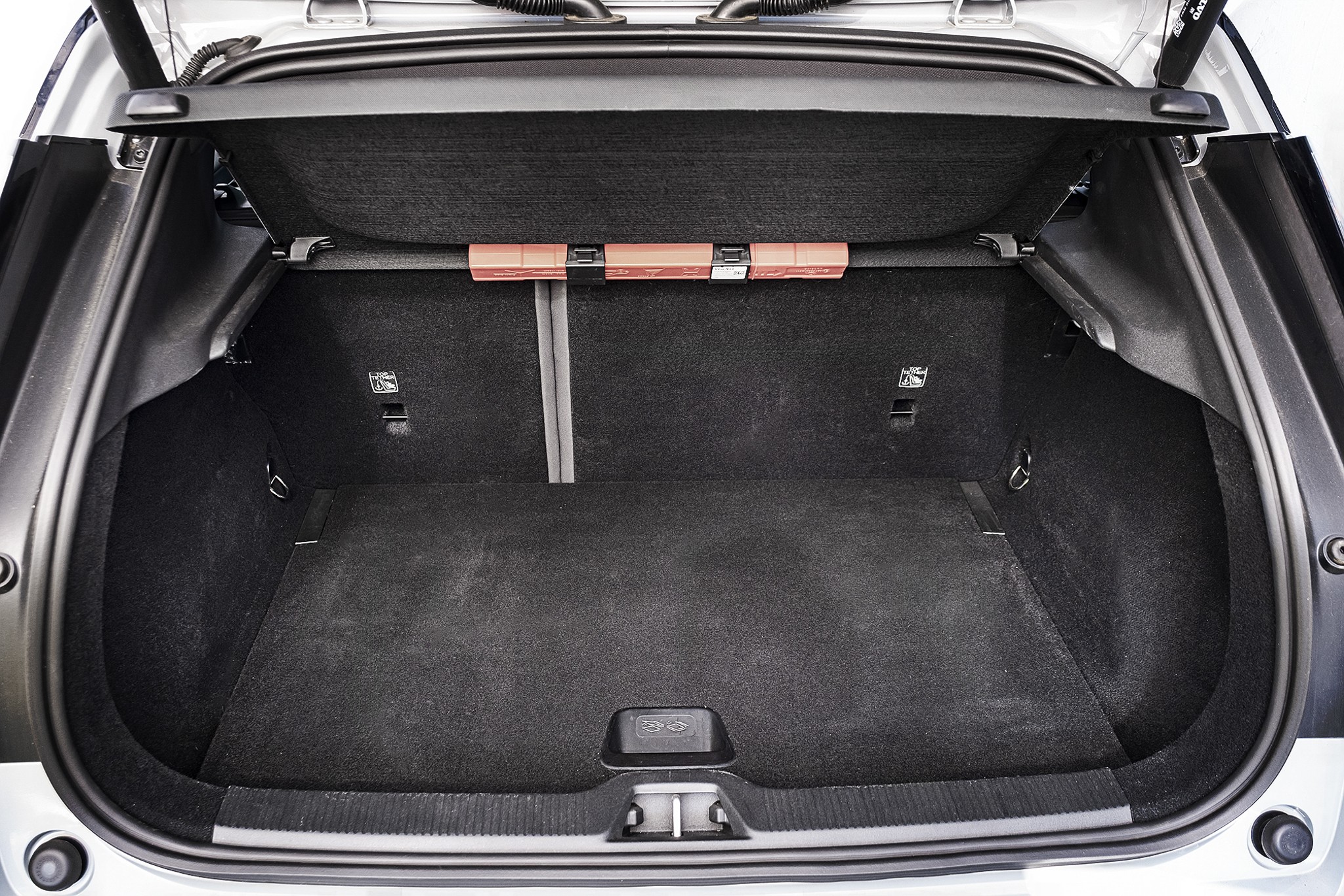 Porta-malas de carro hatch aberto, forrado com carpete cinza escuro, sem objetos. H&aacute; uma barra transversal com tr&ecirc;s refletores laranja na parte superior e ganchos laterais para fixa&ccedil;&atilde;o de carga