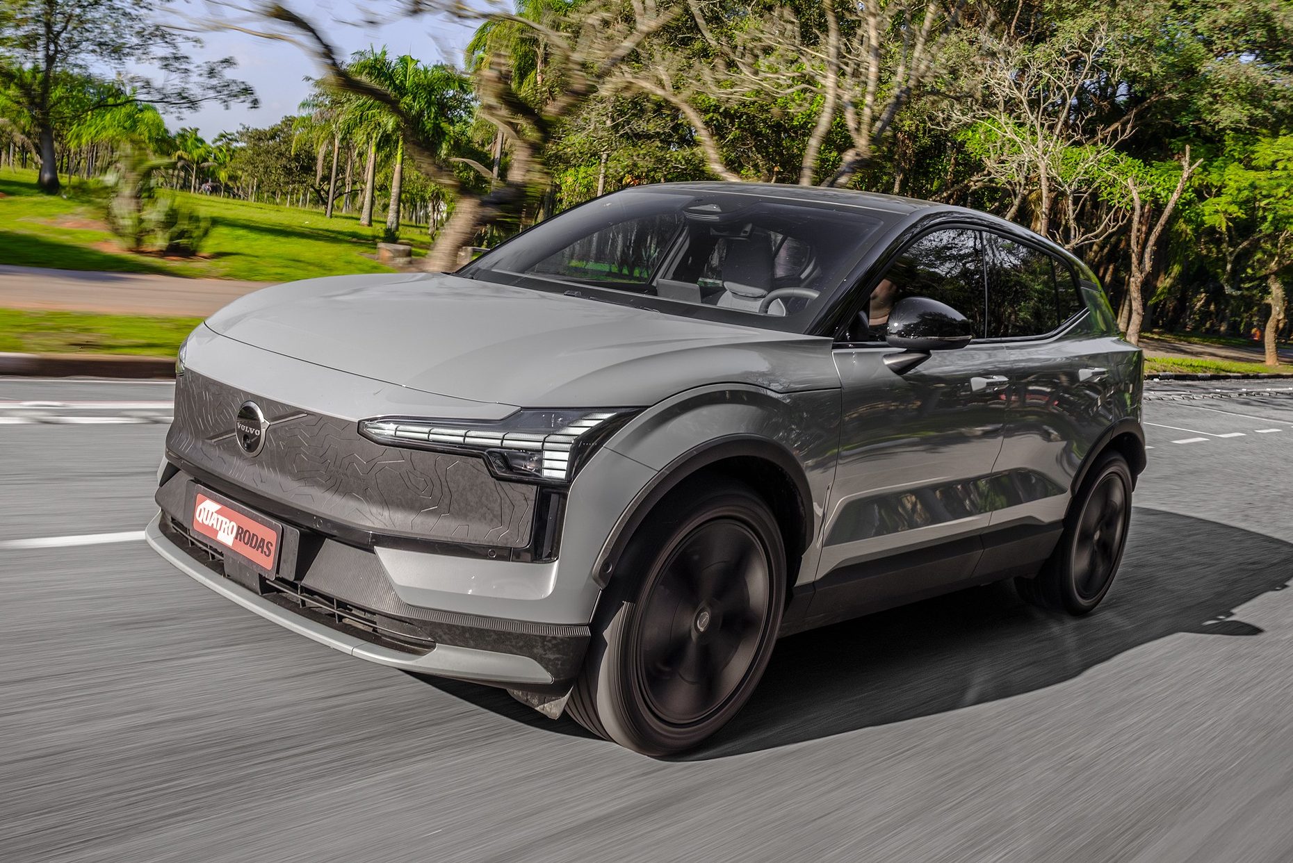 Carro Volvo EX30 cinza claro em movimento, visto de frente e lateral, com rodas escuras e far&oacute;is em formato de T. O ve&iacute;culo est&aacute; em uma rua asfaltada, com &aacute;rvores e gramado ao fundo, sob c&eacute;u azul