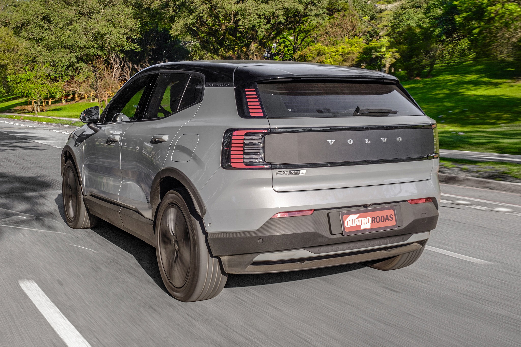 SUV Volvo EX30 cinza claro com teto preto, visto de traseira e lateral, em movimento numa estrada asfaltada com &aacute;rvores verdes ao fundo
