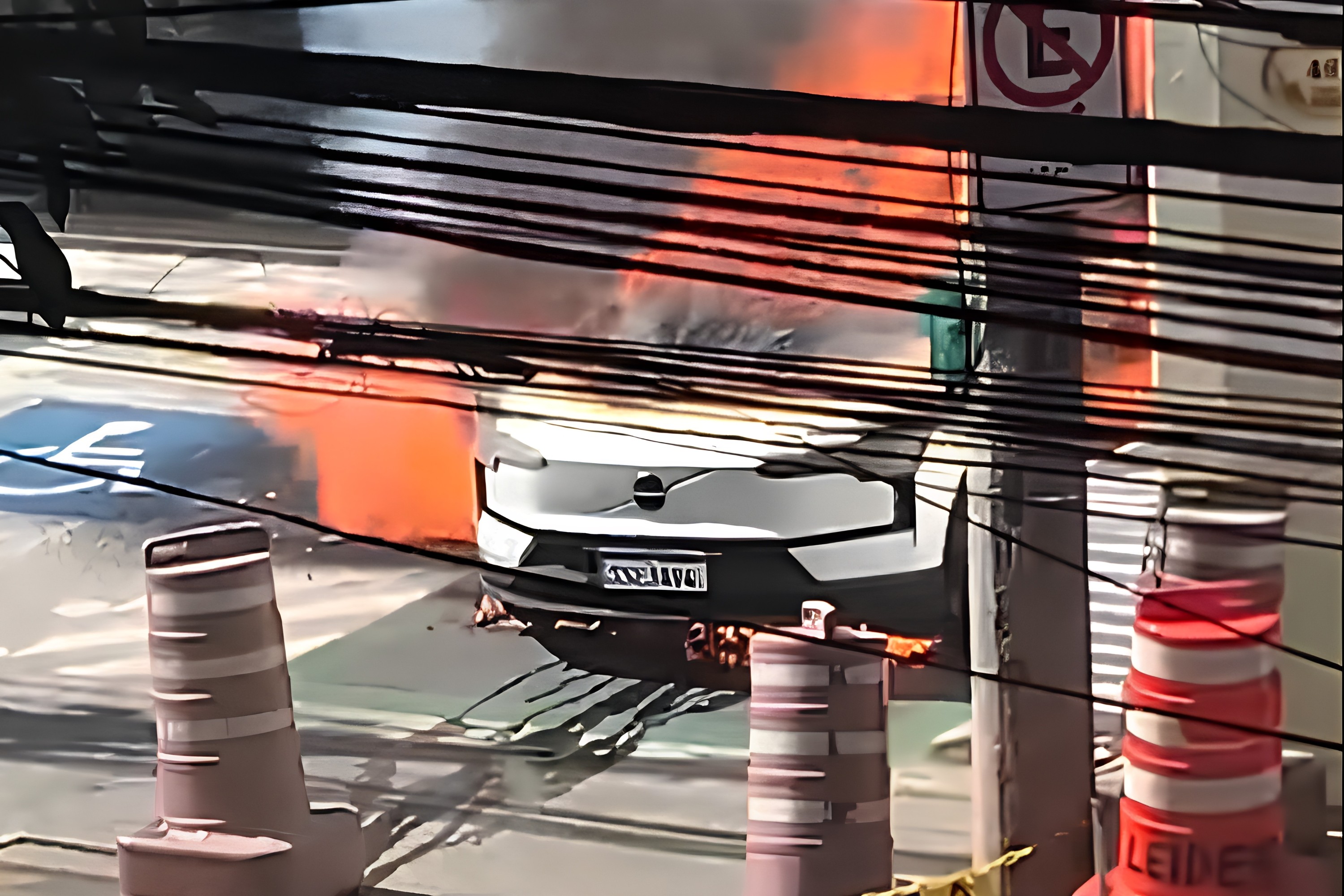 Carro branco em chamas na rua, com fuma&ccedil;a laranja e preta subindo. Fios el&eacute;tricos cruzam a imagem em primeiro plano, e cones de tr&acirc;nsito est&atilde;o vis&iacute;veis. Um poste com placa de proibido estacionar aparece &agrave; direita