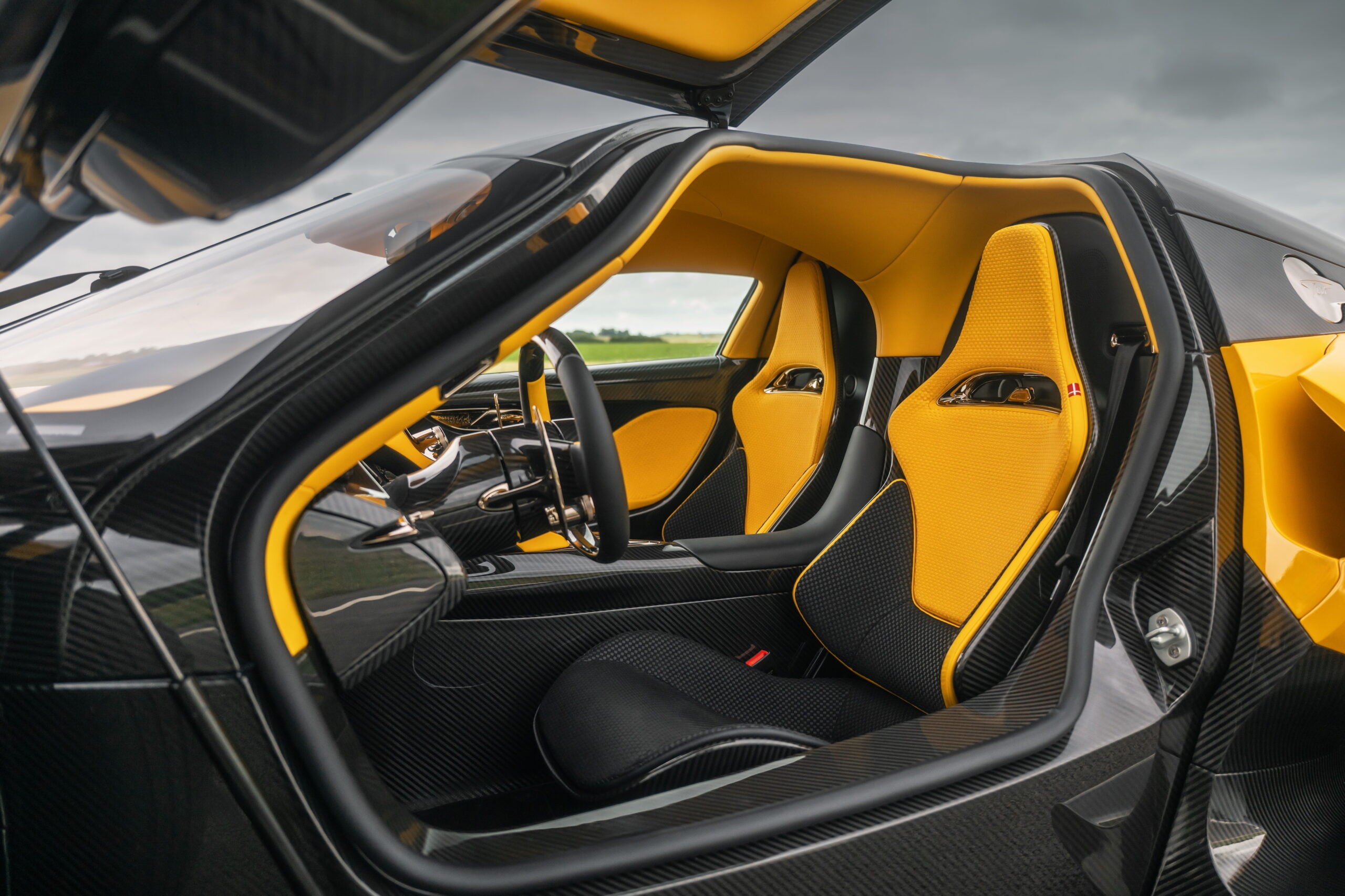 Interior de um carro esportivo de luxo, com a porta aberta revelando bancos esportivos pretos e amarelos, painel e volante pretos com detalhes amarelos, e acabamento em fibra de carbono. O teto tamb&eacute;m &eacute; amarelo.