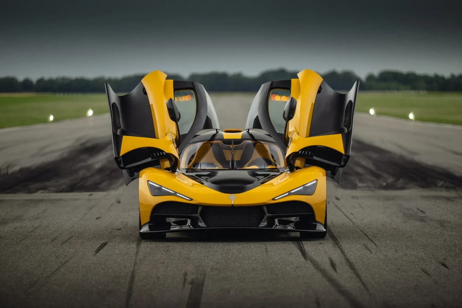 Carro esportivo amarelo e preto com portas abertas para cima, parado em uma pista de asfalto com marcas de pneu, sob um céu nublado