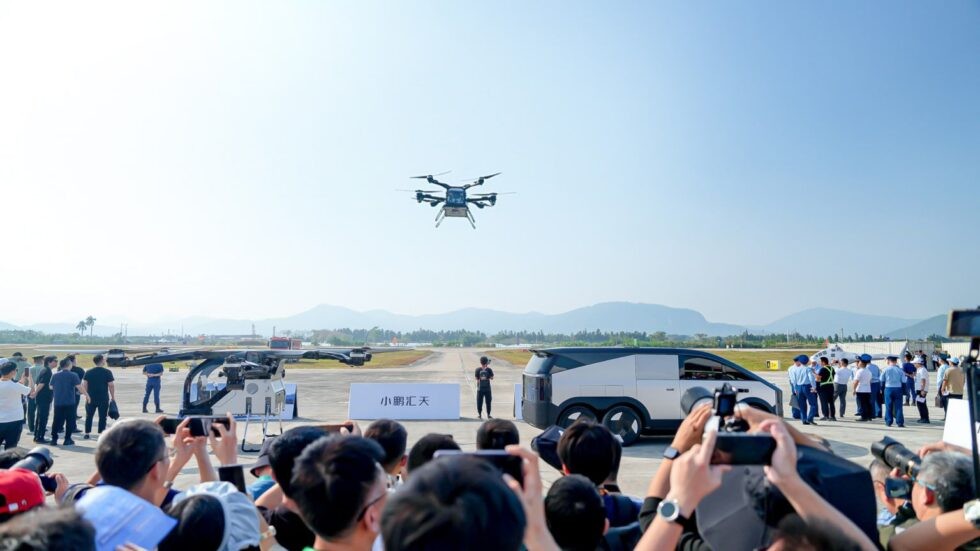 Um drone de oito h&eacute;lices paira no ar, observado por uma multid&atilde;o em um evento ao ar livre. Pessoas filmam com celulares, e um ve&iacute;culo futurista branco est&aacute; &agrave; direita. Montanhas distantes e c&eacute;u azul claro comp&otilde;em o cen&aacute;rio