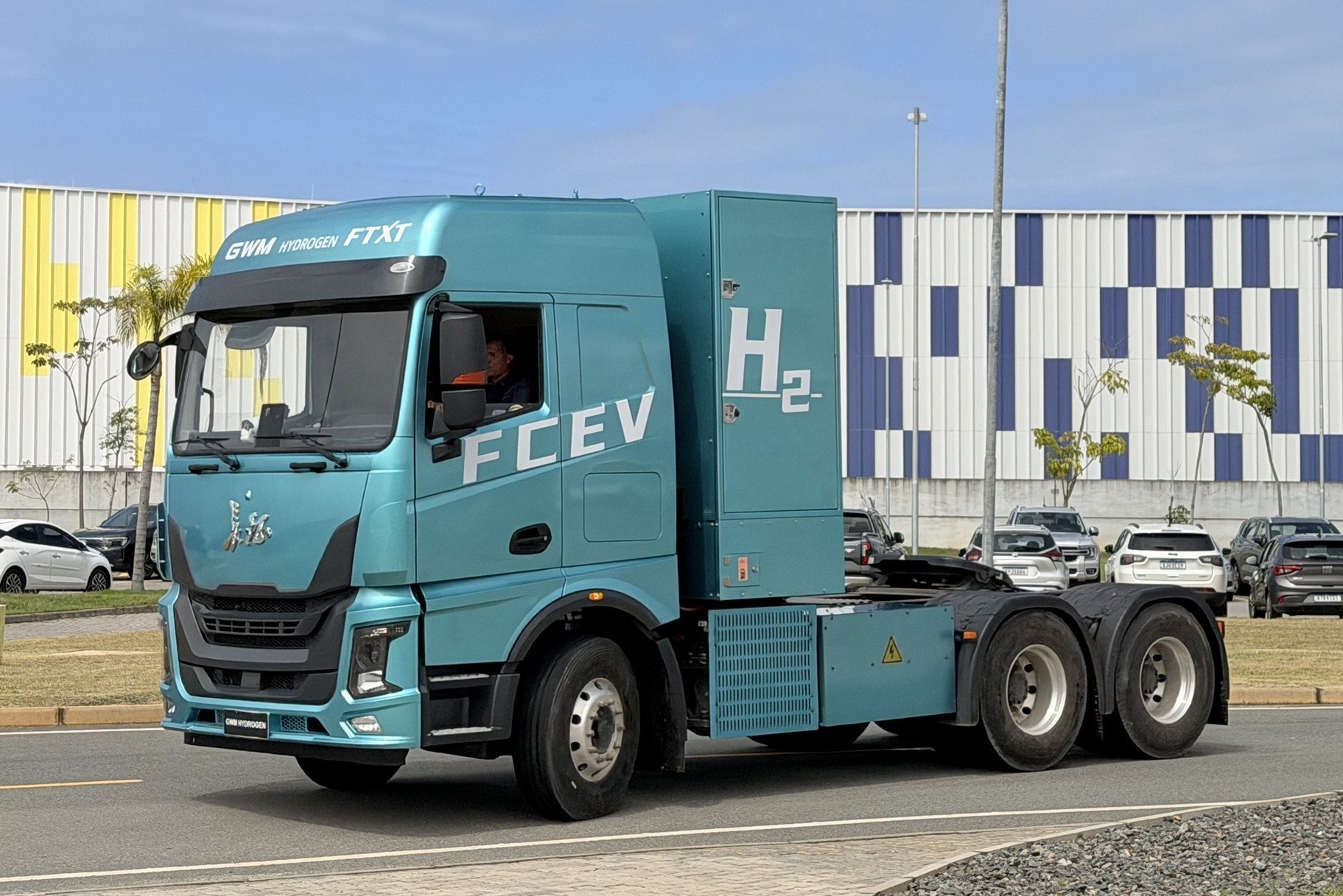 Caminh&atilde;o azul-claro com as inscri&ccedil;&otilde;es FCEV e H2 na lateral, indicando ser movido a hidrog&ecirc;nio. Um motorista est&aacute; vis&iacute;vel na cabine. O ve&iacute;culo est&aacute; em uma via, com um estacionamento e um pr&eacute;dio com fachada azul e branca ao fundo