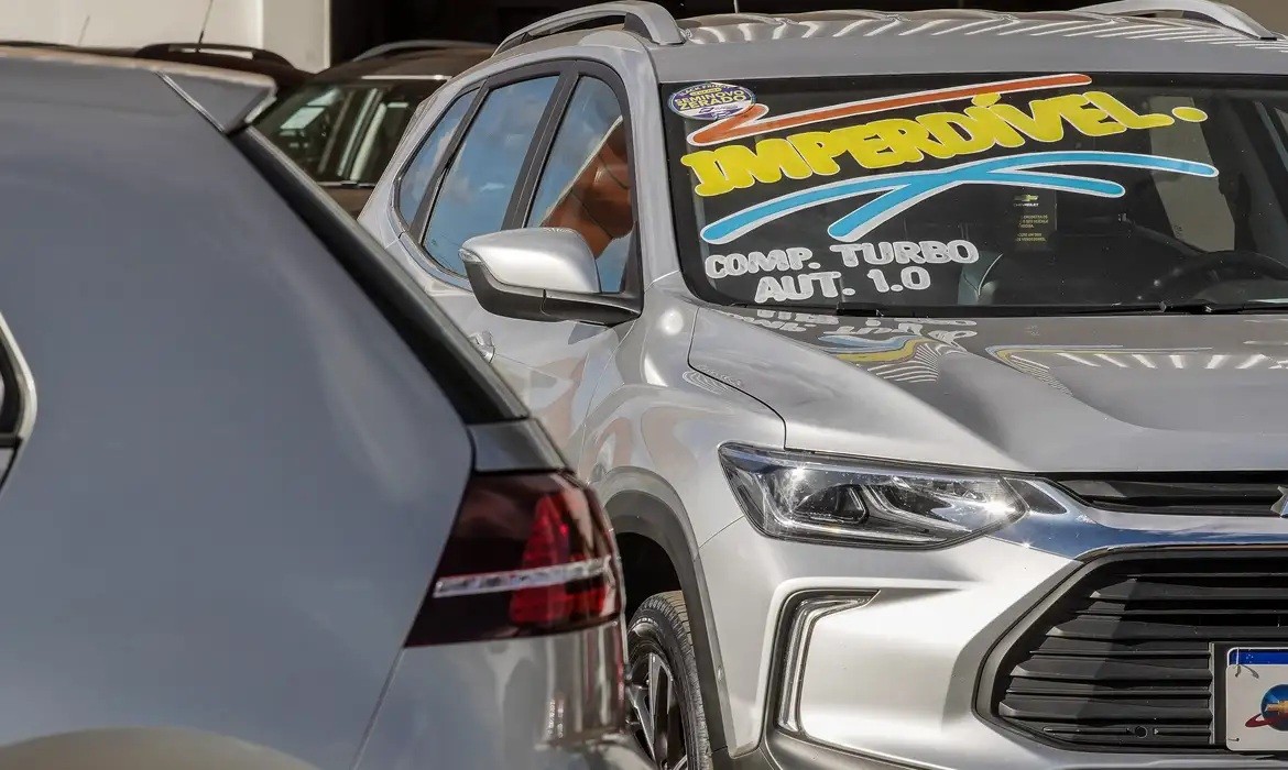 Dois carros prateados estacionados, o da direita &eacute; um SUV com um adesivo no para-brisa escrito IMPERD&Iacute;VEL em amarelo e COMP. TURBO AUT. 1.0 em branco