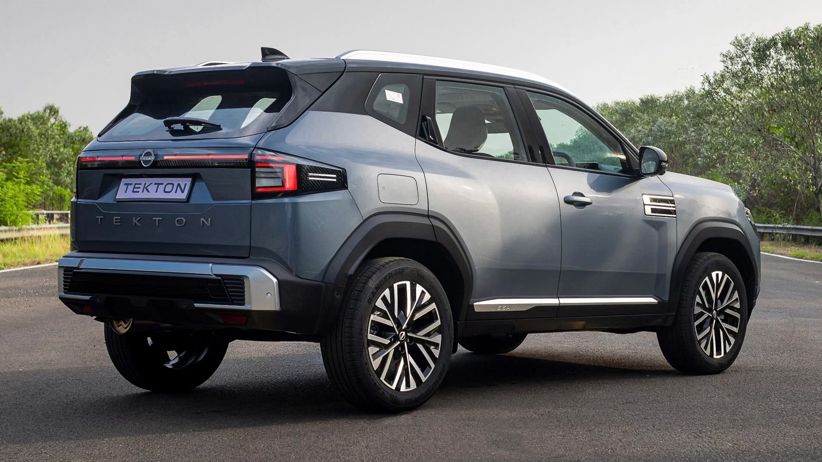 SUV Tekton cinza-azulado estacionado em uma estrada asfaltada, visto de traseira e lateral. Possui teto branco, rack de teto, rodas com detalhes em preto e prata, e o nome TEKTON na tampa do porta-malas. Ao fundo, vegeta&ccedil;&atilde;o e c&eacute;u claro