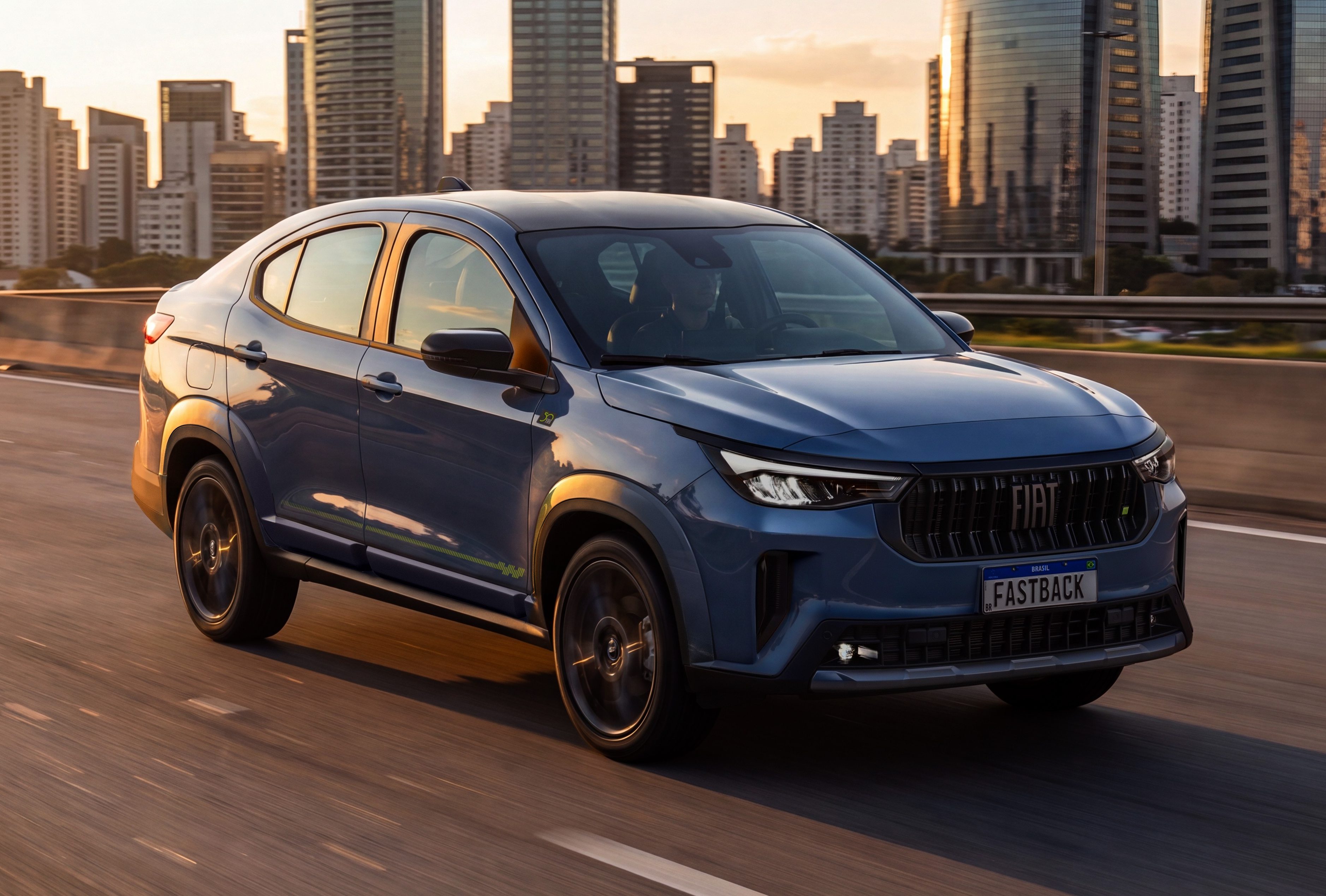 Carro azul escuro, modelo Fiat Fastback, em movimento numa rodovia com pr&eacute;dios altos ao fundo, sob o p&ocirc;r do sol