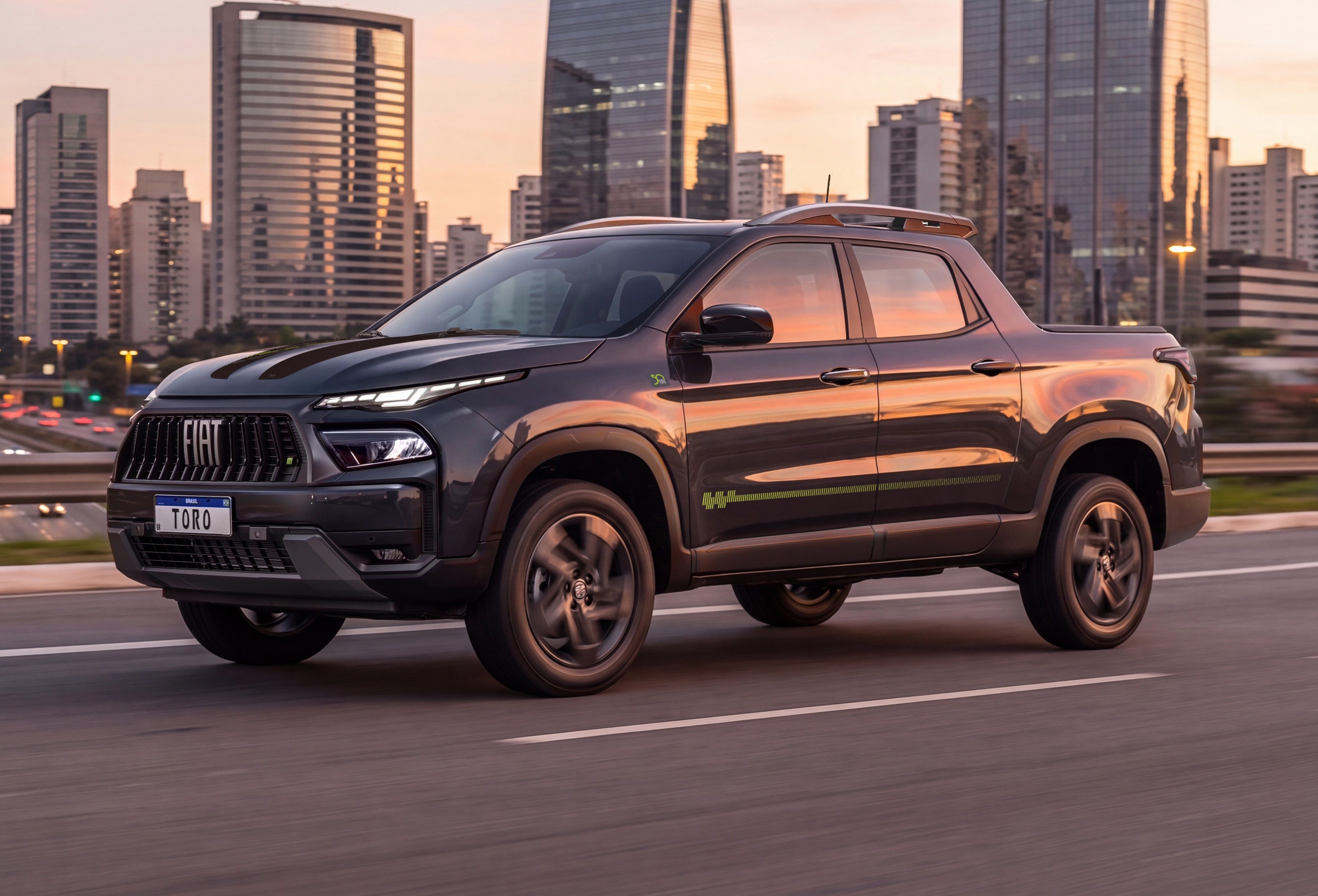 Picape Fiat Toro cinza escura com detalhes verdes na lateral, em movimento numa rodovia. Ao fundo, pr&eacute;dios altos de uma cidade e o c&eacute;u alaranjado do p&ocirc;r do sol. Carros com far&oacute;is acesos na pista oposta