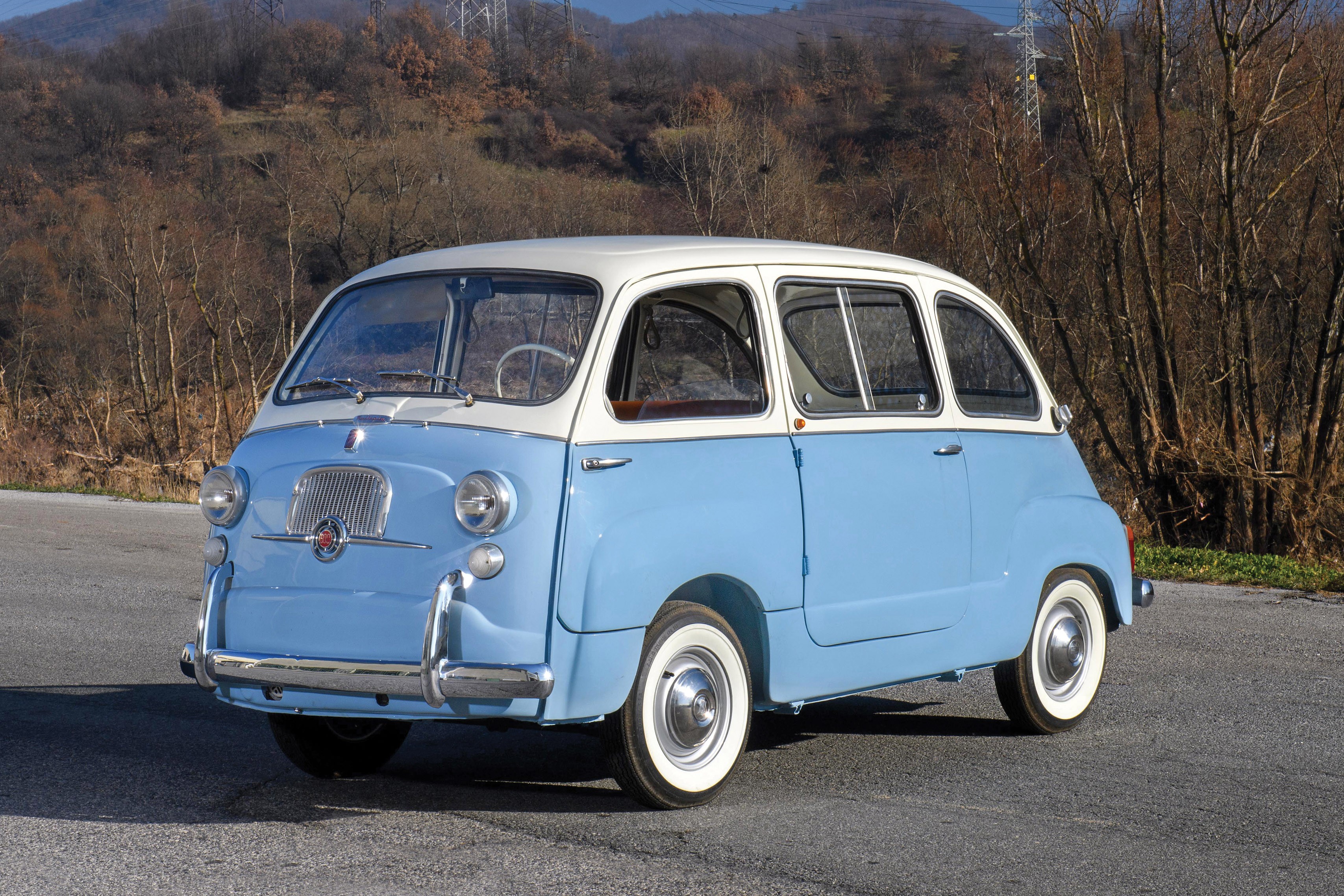 Uma van Fiat 600 Multipla azul claro e branca, estilo retr&ocirc;, estacionada em uma estrada asfaltada com &aacute;rvores secas e montanhas ao fundo