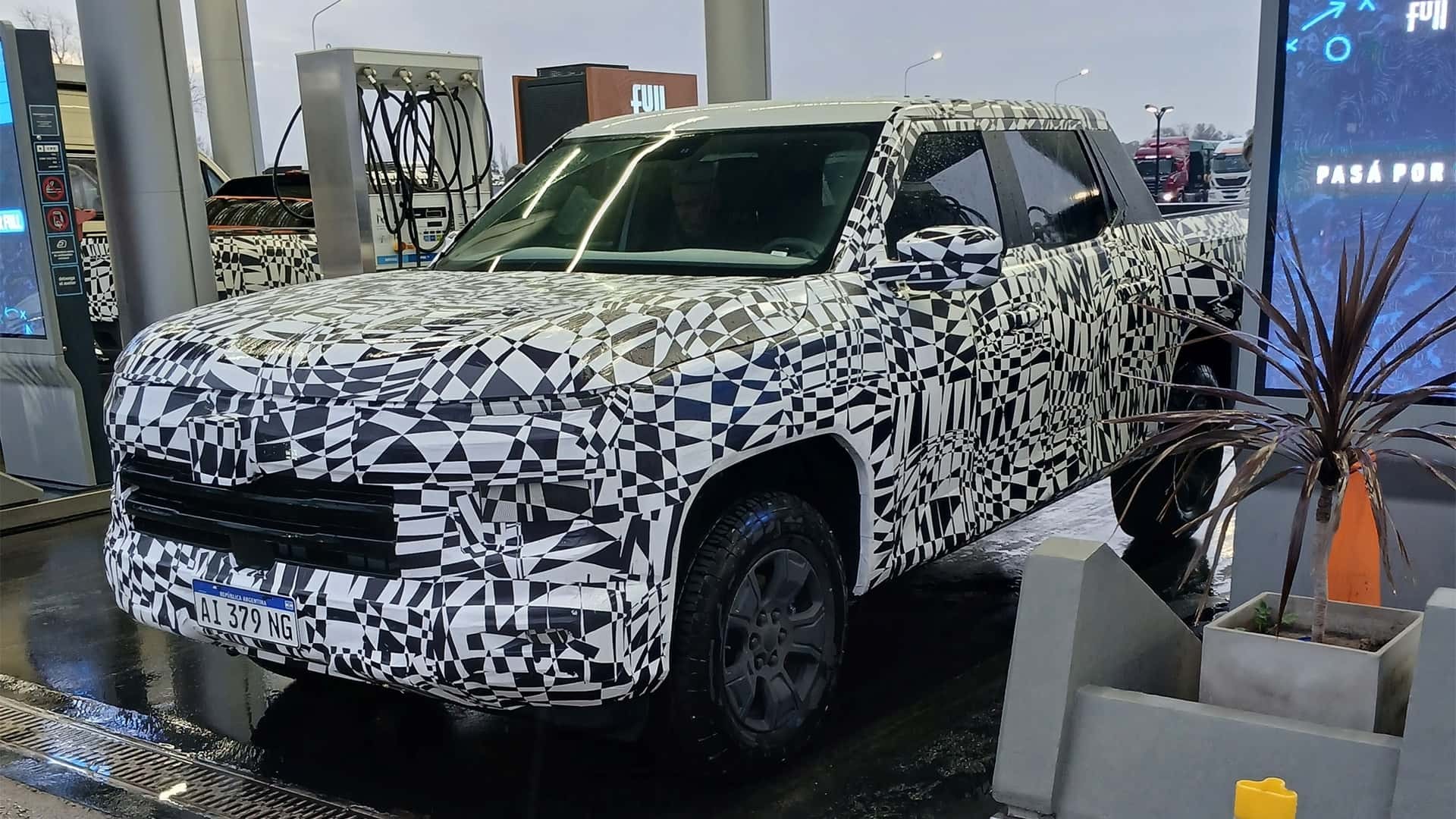 Picape camuflada com adesivos pretos e brancos em padr&atilde;o geom&eacute;trico, estacionada em posto de combust&iacute;vel. A placa frontal exibe AL 379 NG. Ao fundo, bombas de gasolina e um painel digital com a frase PAS&Aacute; POR. &Agrave; direita, um vaso com planta de folhas escuras