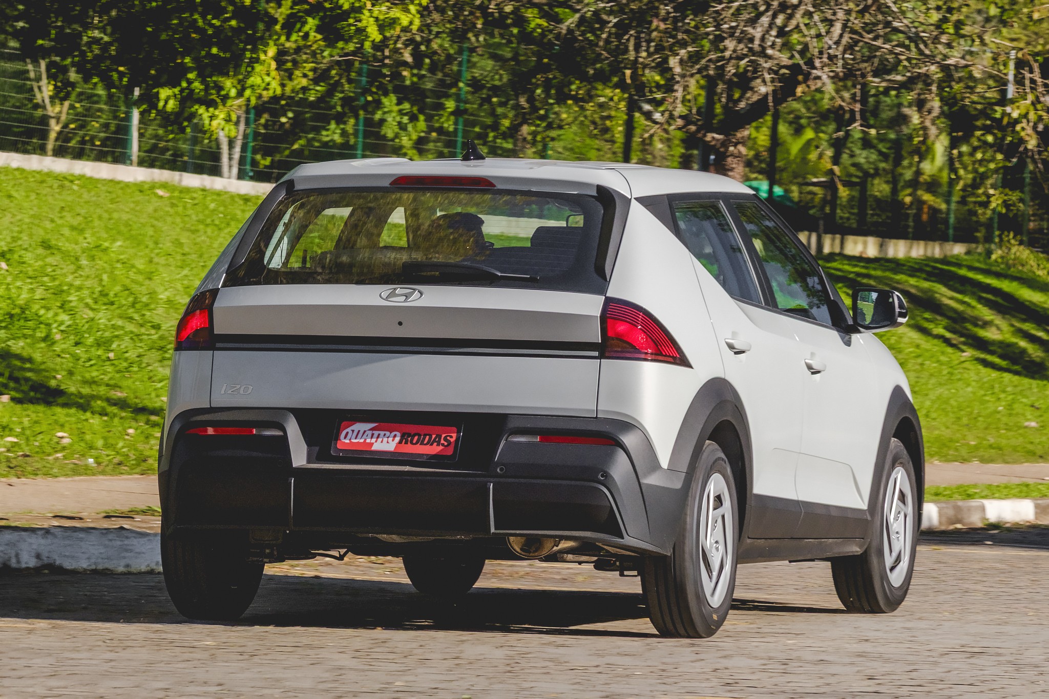 Traseira de um carro Hyundai branco, modelo SUV compacto, estacionado em uma rua pavimentada. O veículo possui lanternas vermelhas triangulares, para-choque preto e detalhes em cinza. Ao fundo, gramado verde e árvores frondosas sob luz solar