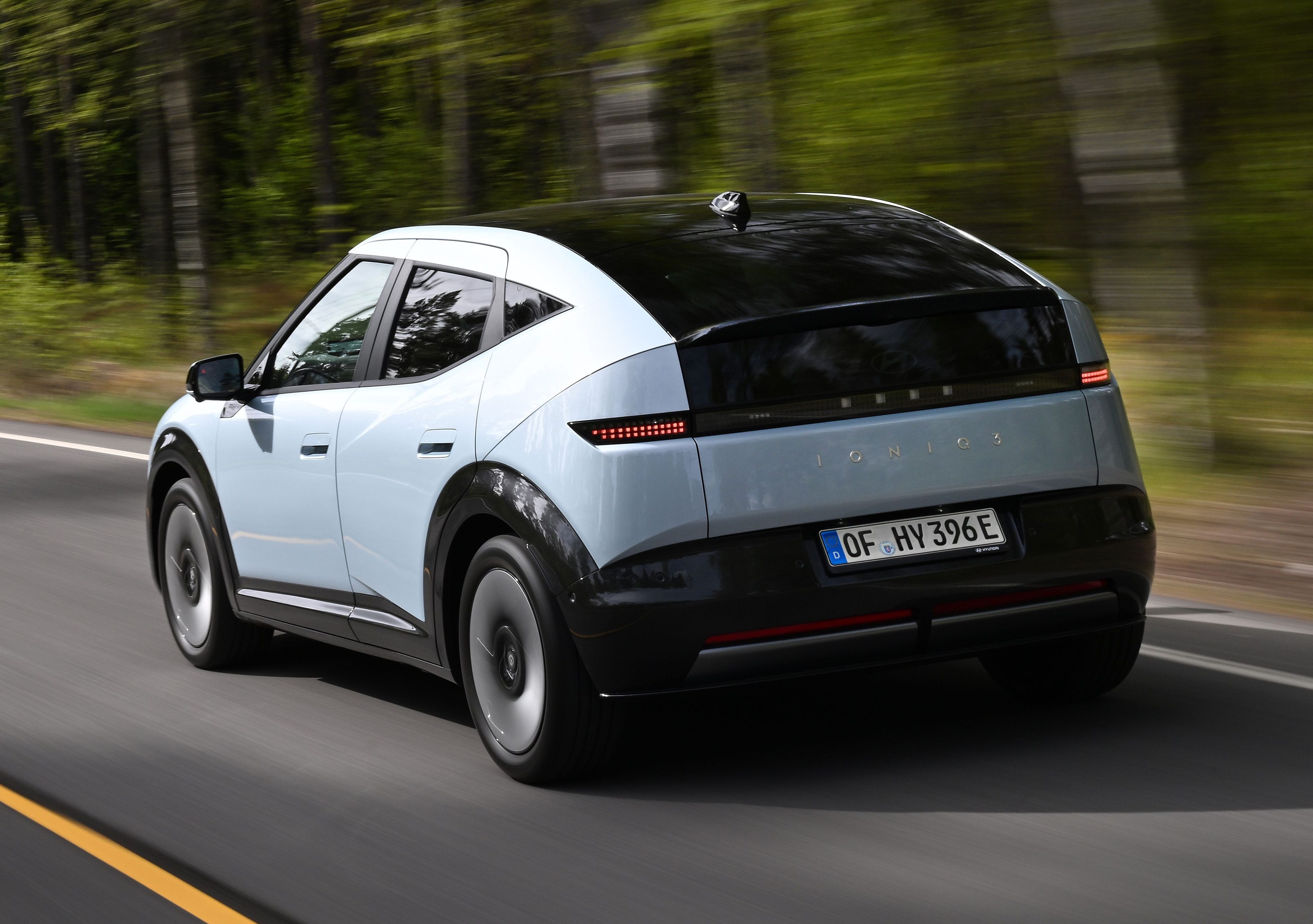 Carro el&eacute;trico branco e preto em movimento, visto por tr&aacute;s, em uma estrada asfaltada com faixa amarela e branca, cercada por &aacute;rvores verdes e c&eacute;u nublado