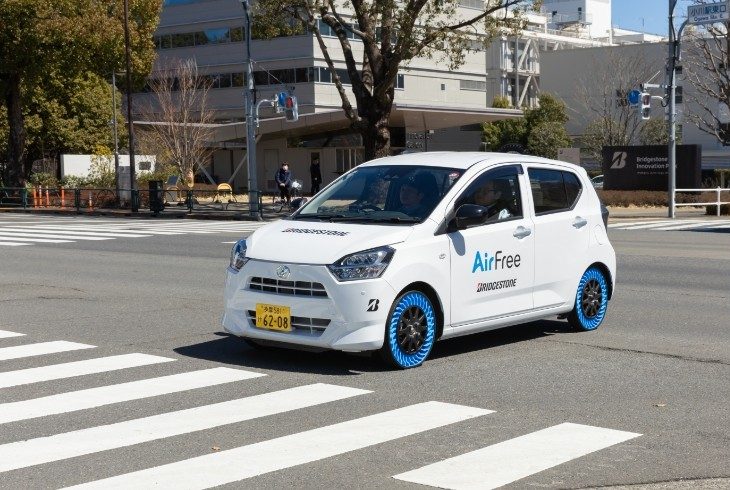 Dois carros brancos id&ecirc;nticos, com pneus azuis sem ar, cruzam uma faixa de pedestres em uma rua urbana ensolarada