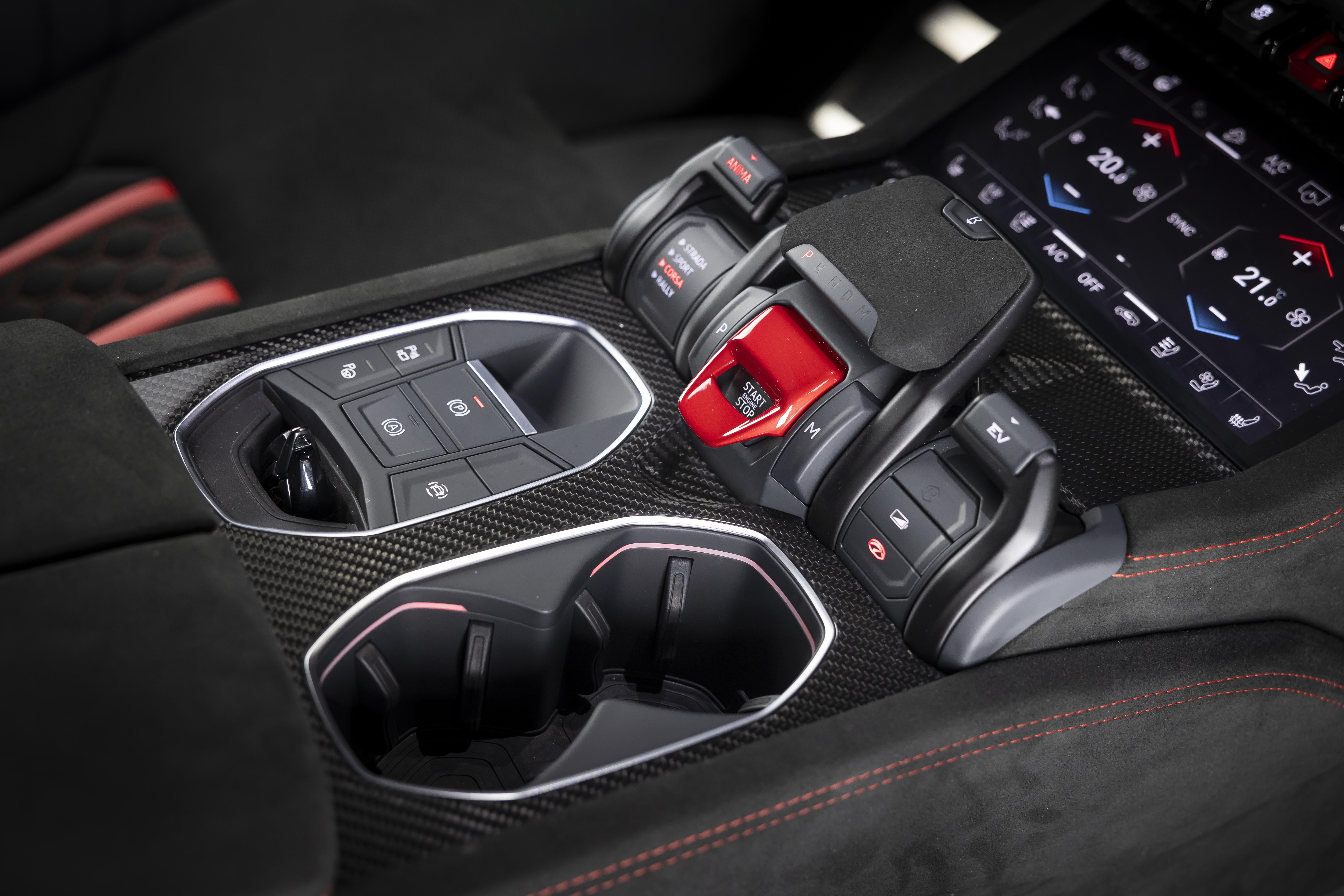Console central de um carro esportivo, com acabamento em fibra de carbono. &Agrave; esquerda, um painel de bot&otilde;es de controle. Ao centro, o bot&atilde;o vermelho de partida do motor, protegido por uma tampa. &Agrave; direita, uma tela digital exibe controles de temperatura. Abaixo, dois porta-copos. O estofamento preto com costura vermelha &eacute; vis&iacute;vel nas laterais